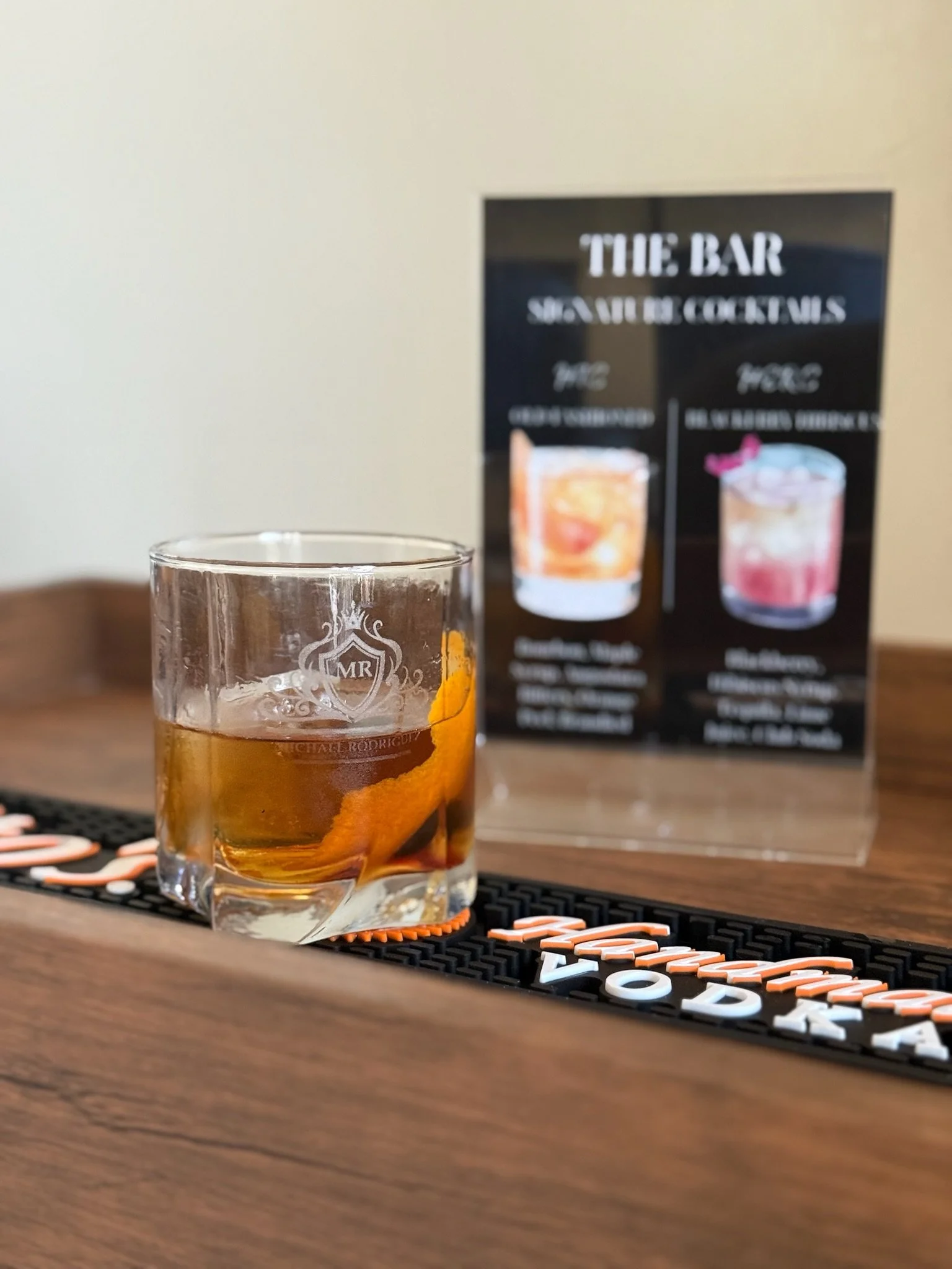 The "his" wedding cocktail in a glass of whiskey on a black bar mat with orange and white lettering, and a menu behind it showing two cocktail options.