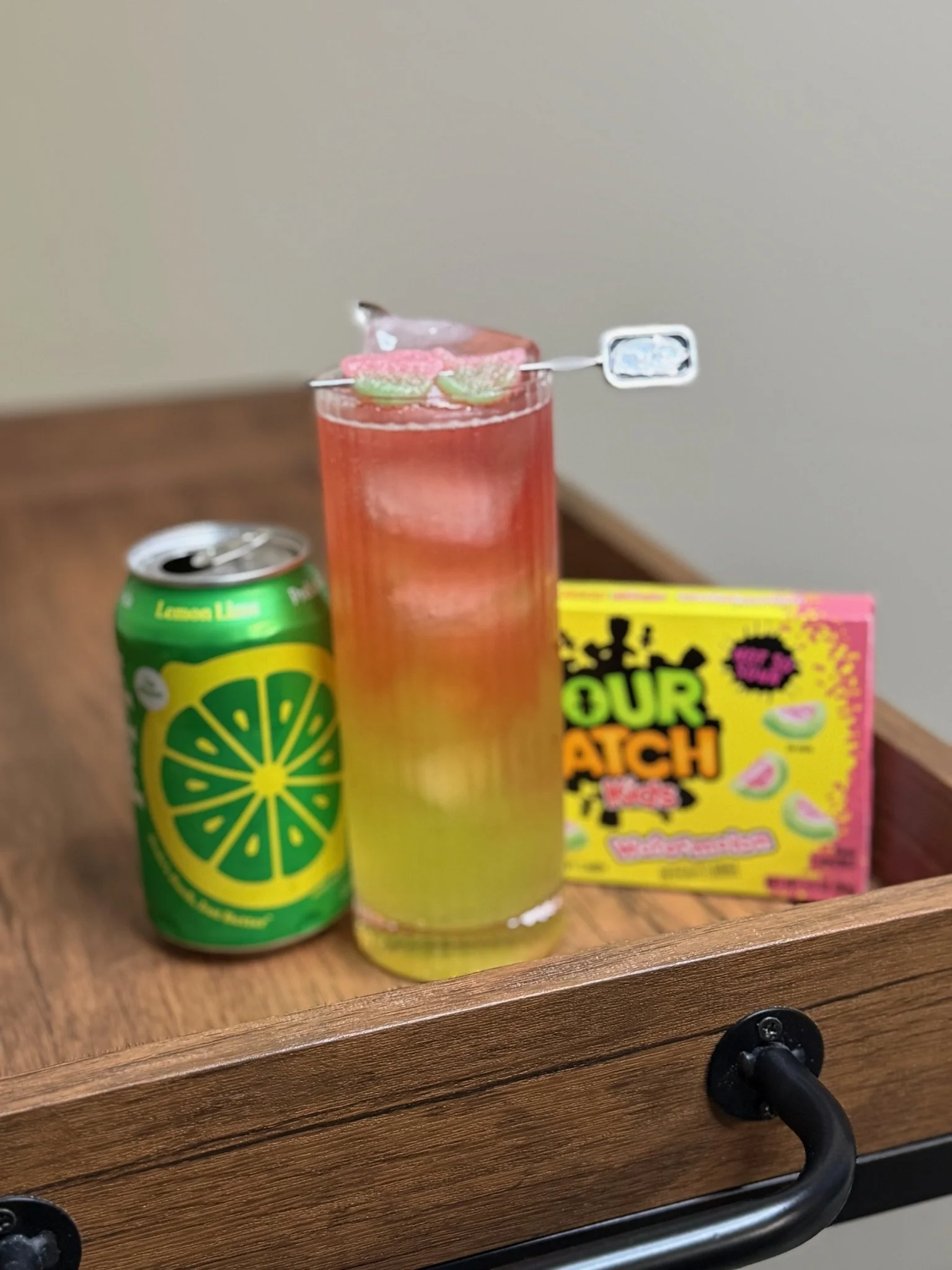 A fun and refreshing signature drink called the "watermelon sugar" made by after two mixology. It has a  can of Sprite Lemon Lime soda, a glass of pink soda with ice and gummy candies, and a box of Warheads sour candy on a wooden surface.