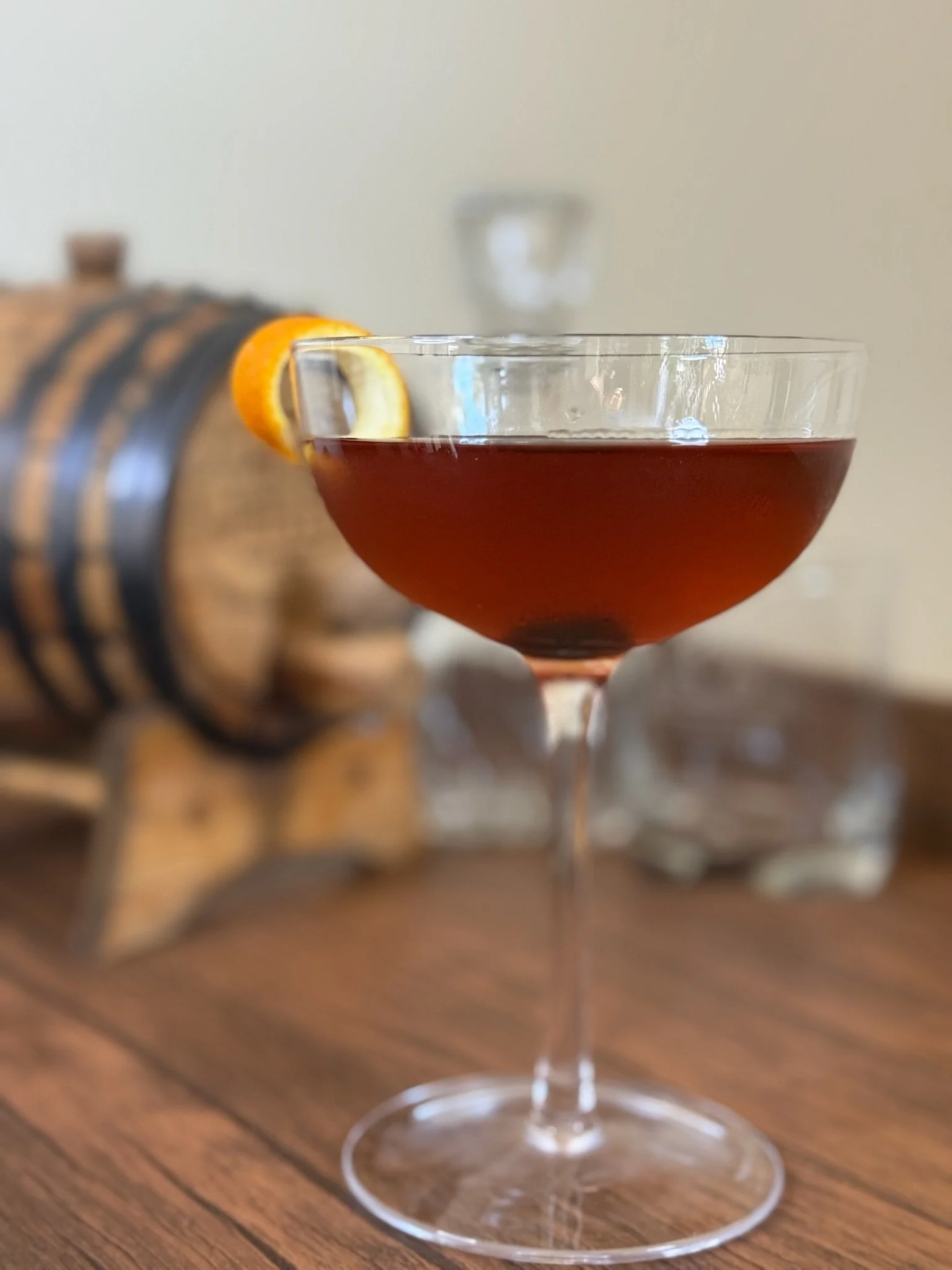 A cocktail glass filled with a dark red beverage, garnished with a twist of orange peel, placed on a wooden surface with a barrel in the background