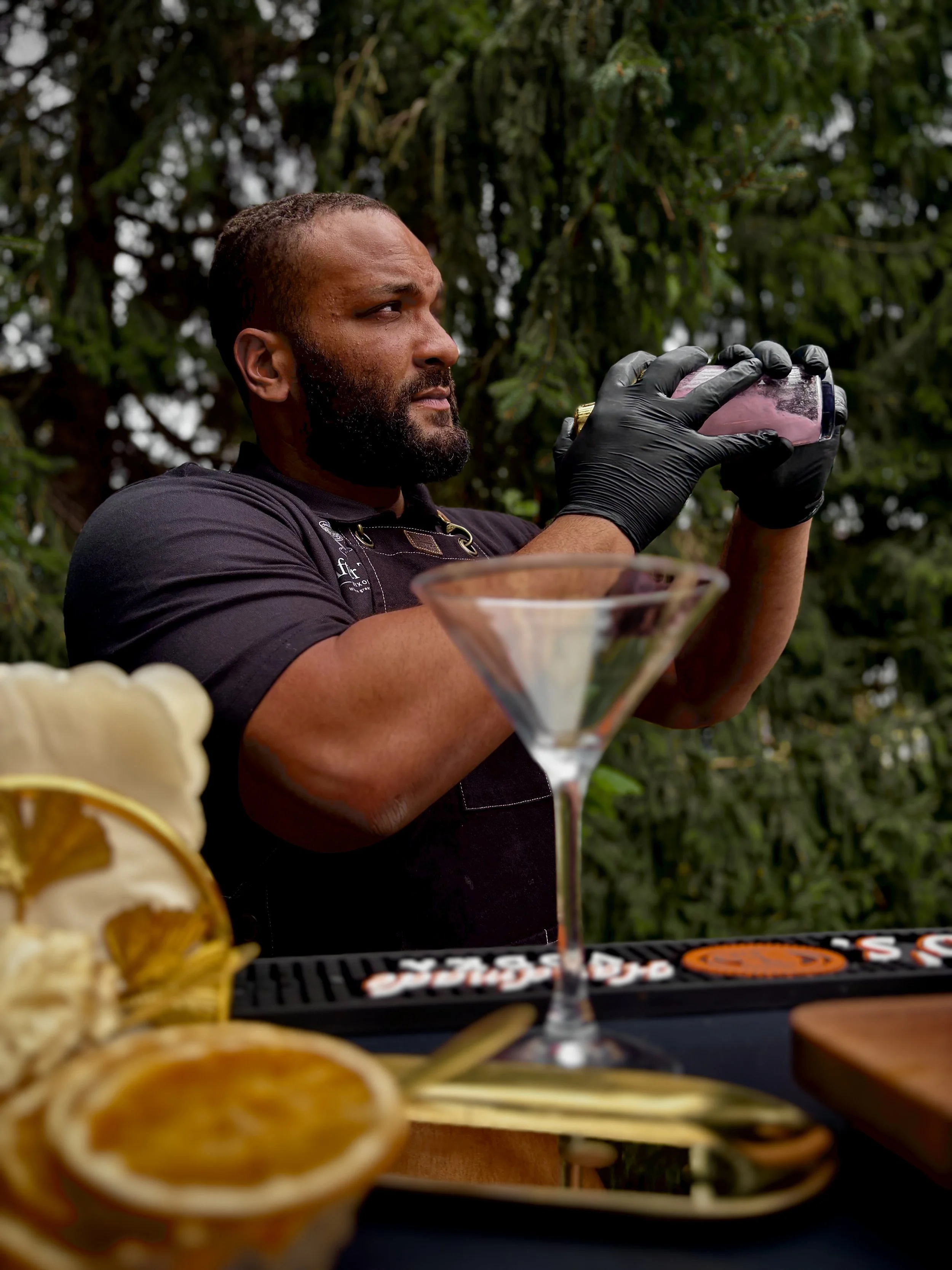 A man with a beard wearing black gloves is shaking a cocktail with a background of trees. In the foreground, there are sliced lemons, a pineapple, and a cocktail glass with a pink drink.