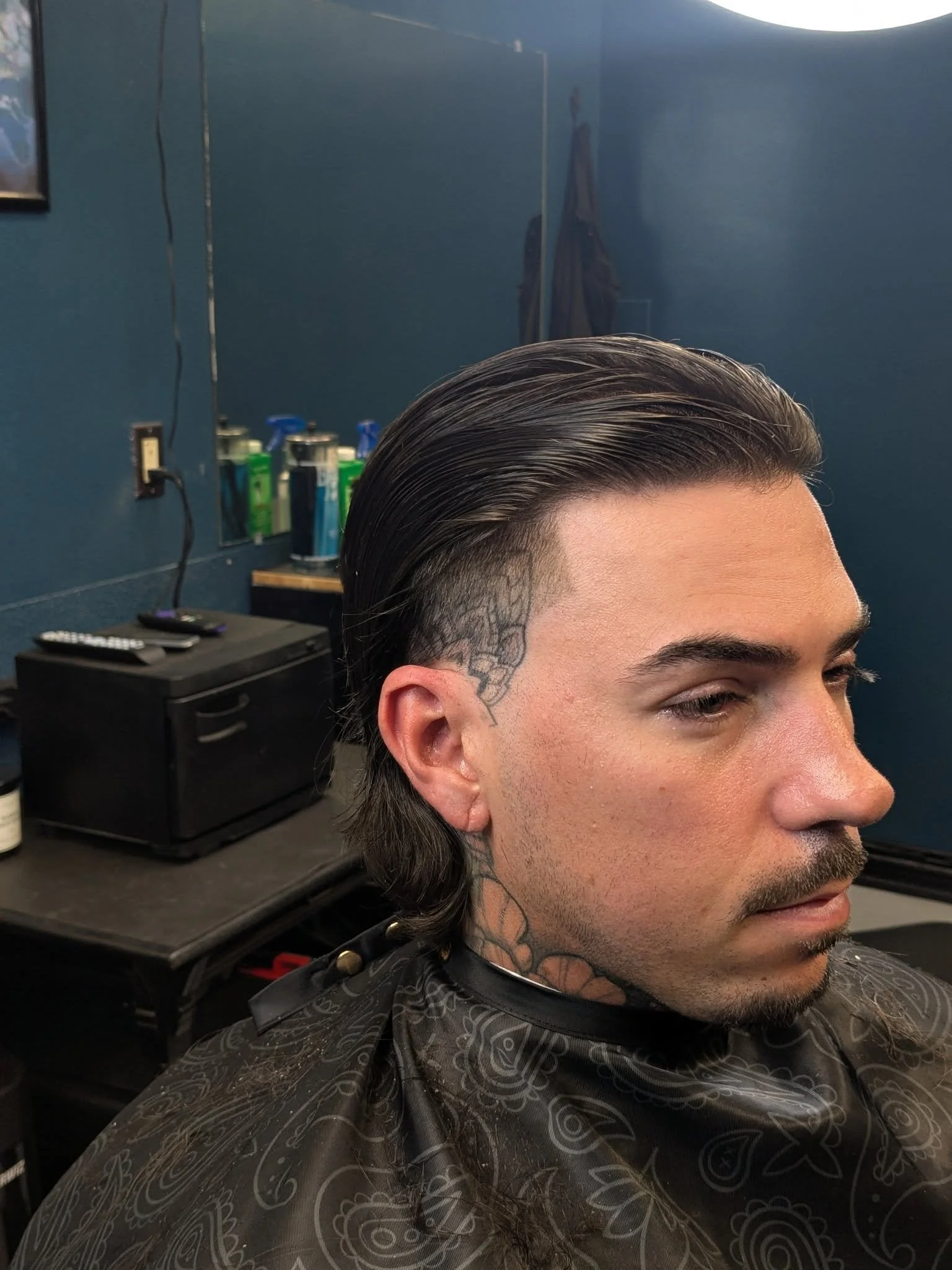 Man with slicked-back hair and tattoos on the side of his head and neck sitting in a barber shop chair.