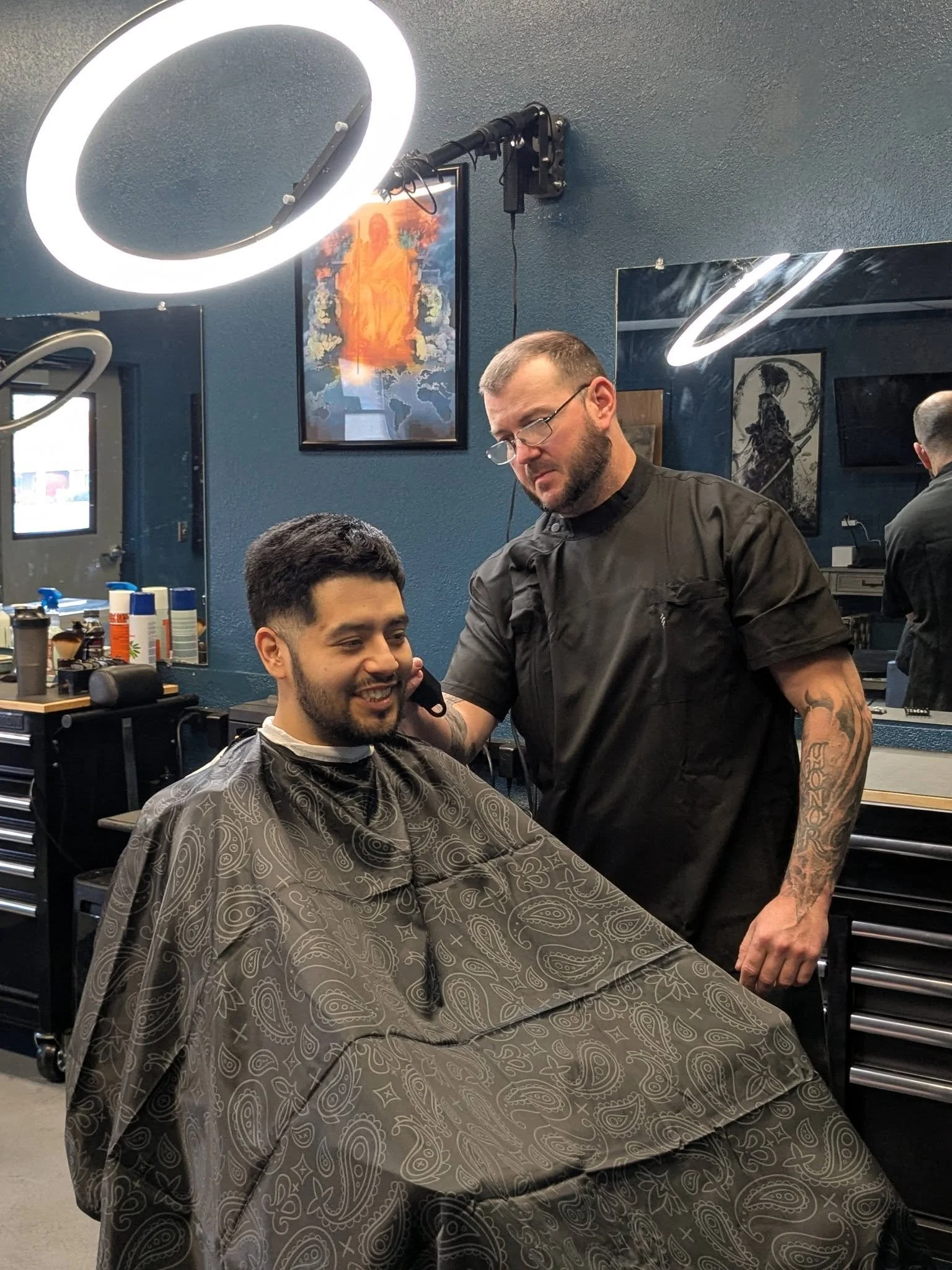 A man getting a haircut in a salon, sitting in a chair with a patterned cape, while the barber, wearing glasses and a black shirt, trims his hair.