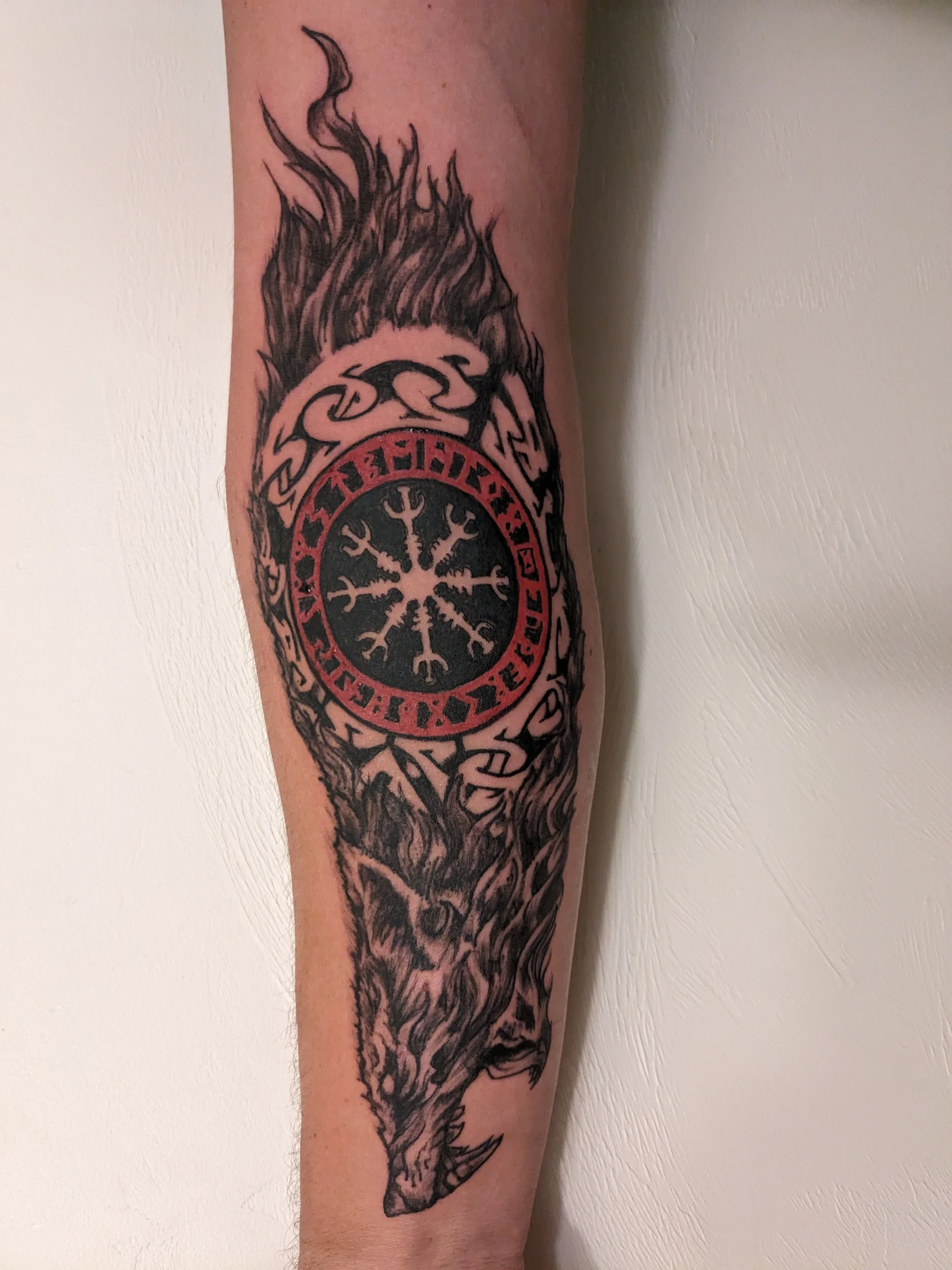 Tattoo of a wolf's head with flames, a central circular symbol with a snowflake-like design, black and red in color, on a person's forearm.