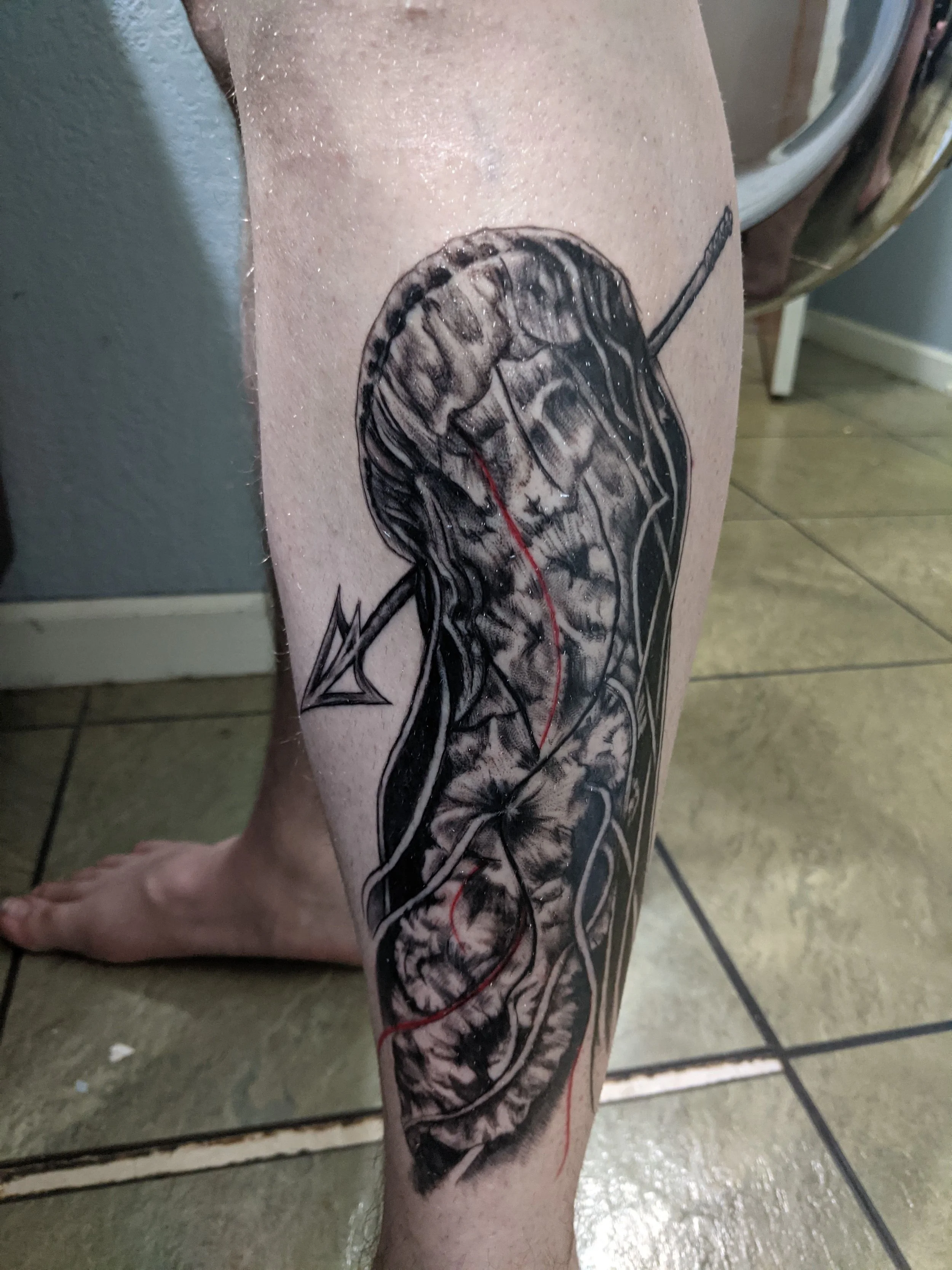 A black and gray tattoo of a human brain with a skull overlay, featuring intricate shading and line work, with red lines running through the design.