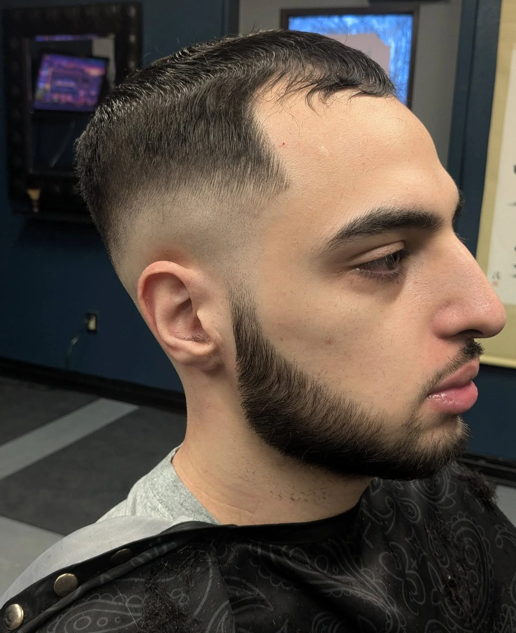 Side profile of a young man with a freshly cut hairstyle and beard, sitting in a barbershop.