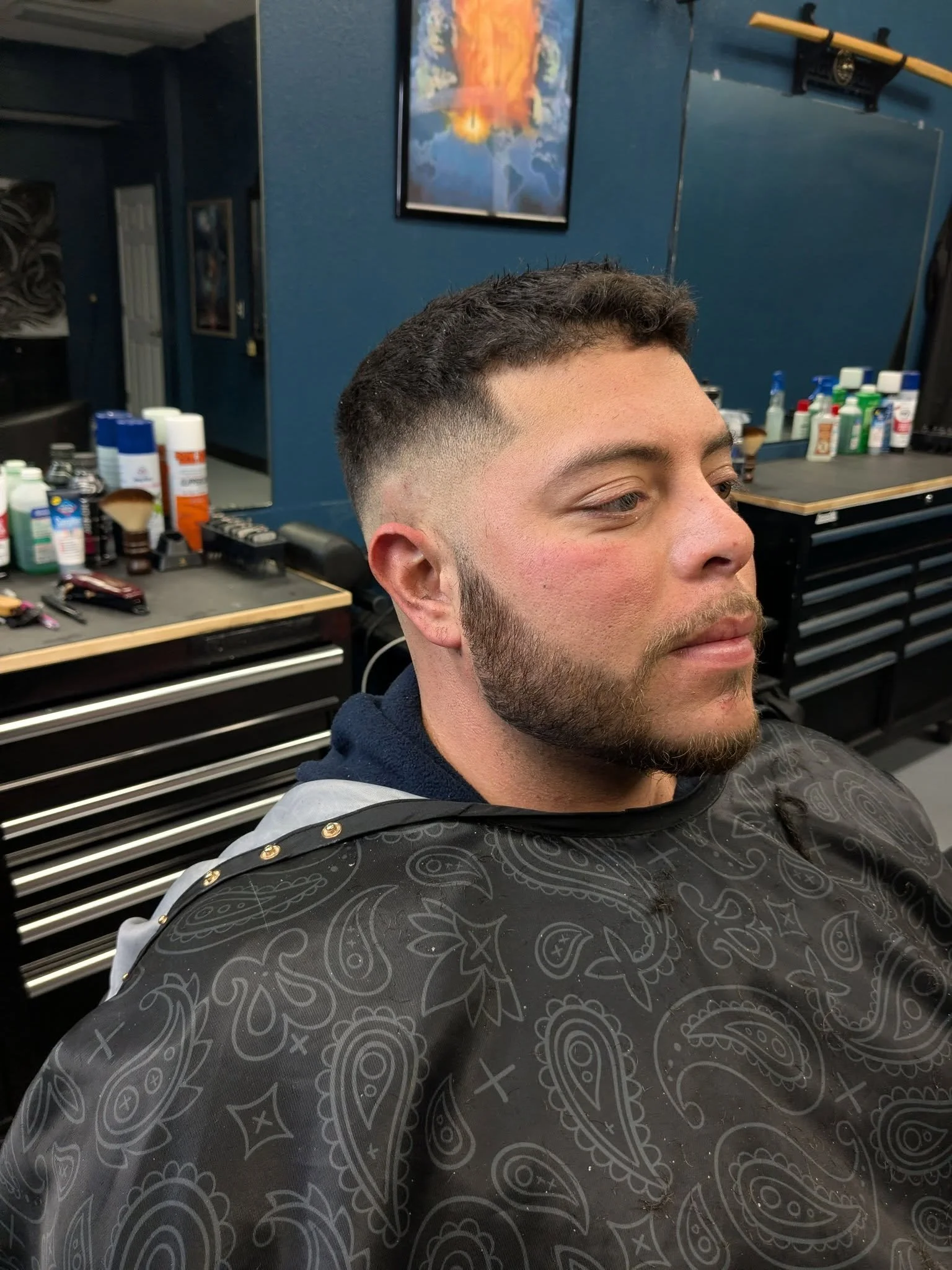 A man with a fresh fade haircut, facial hair, and closed eyes sitting in a barbershop chair. The background shows barber supplies on a workstation, a blue wall with a colorful abstract painting, and a hanging mirror.
