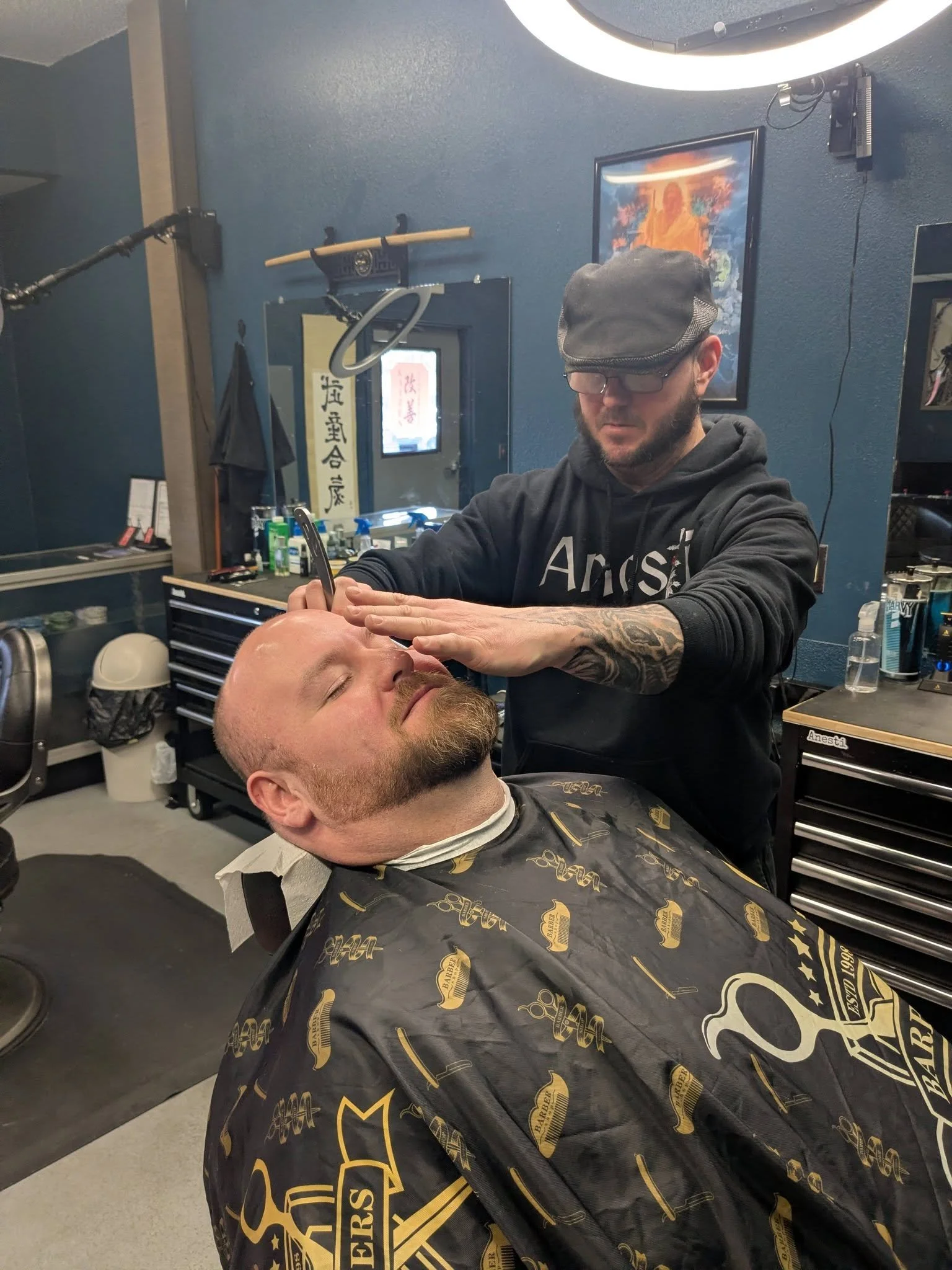 A man is sitting in a barber chair with his eyes closed, getting a haircut or beard grooming. The barber, standing behind him, is using a tool near the man's face. The barber is wearing glasses, a black cap, a black hoodie, and has tattoos on his arms. The scene takes place in a barbershop with black and blue decor, tools, and artwork on the walls.