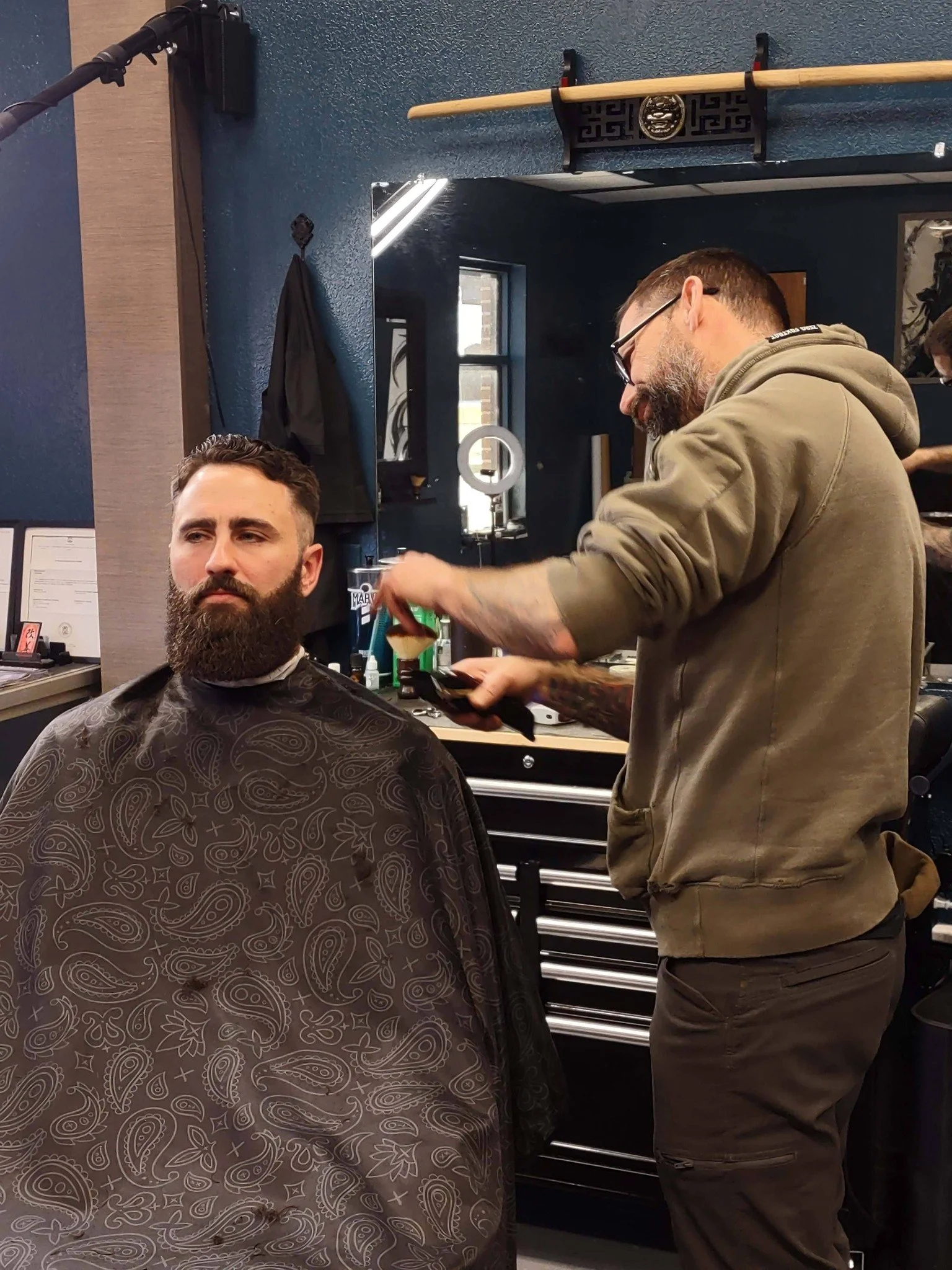 A man getting a haircut in a barber shop, seated with a patterned cape around his shoulders, while a barber with glasses and tattoos cuts his hair.