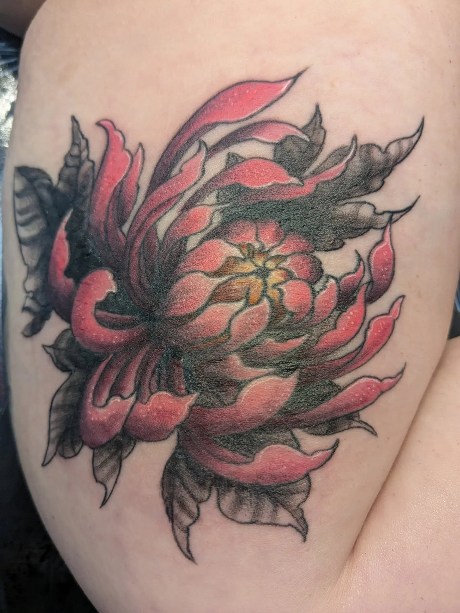 Pink and black floral tattoo on skin featuring a large blooming flower with detailed petals and leaves.