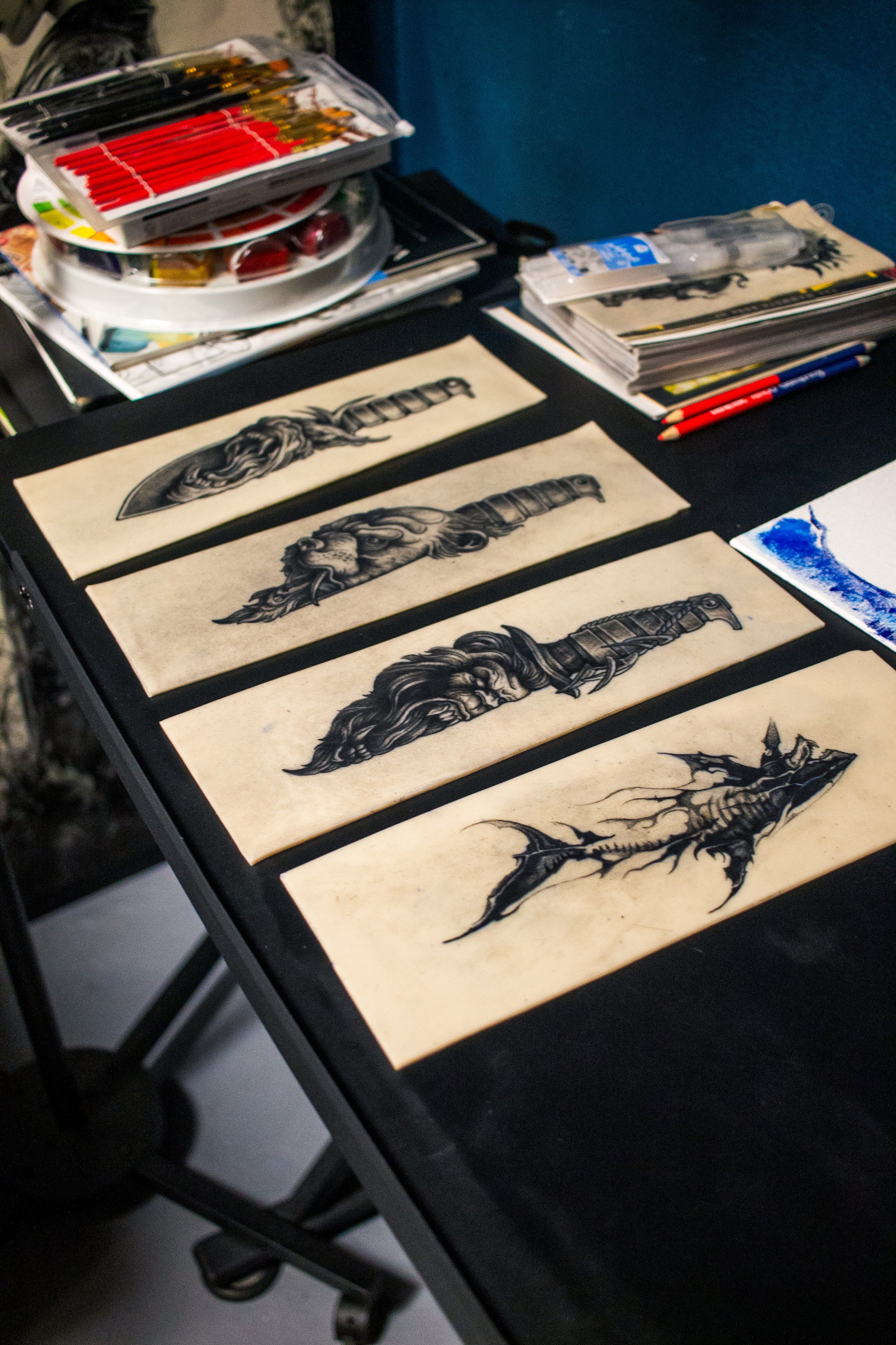 Several sheets of paper with black and gray tattoo designs of fantasy and mythical creatures laid out on a black table, with art supplies and sketches in the background.