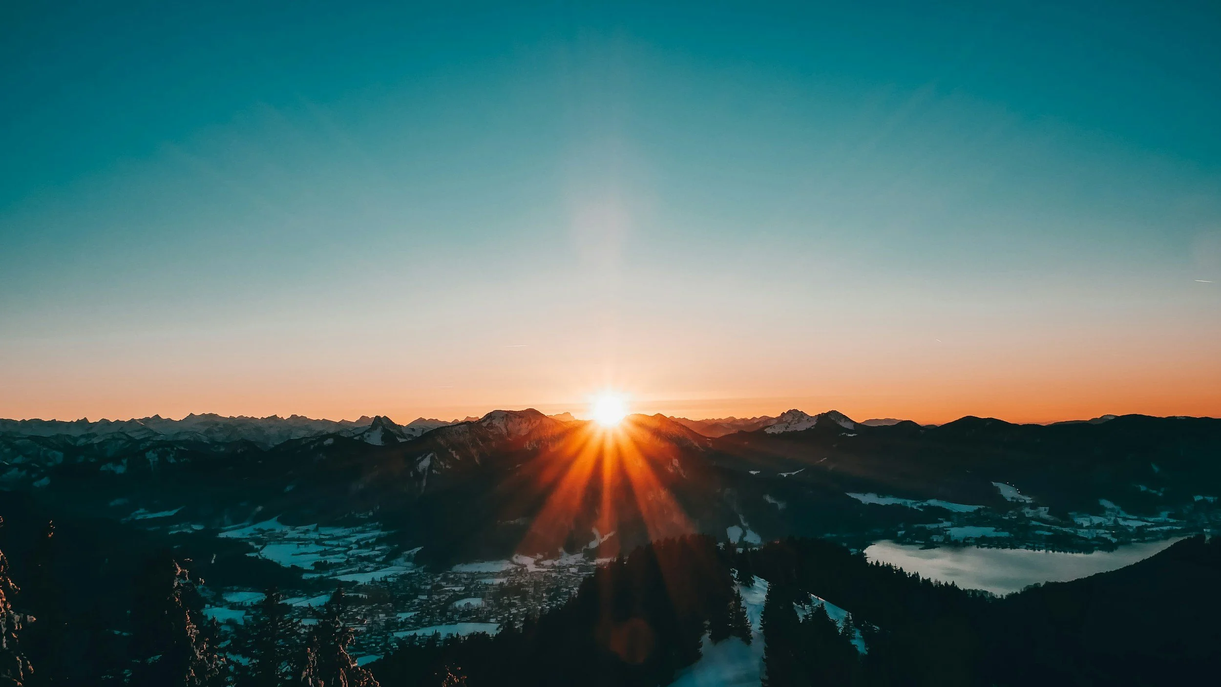 Sunrise over snow-capped mountains and a valley with a lake and trees