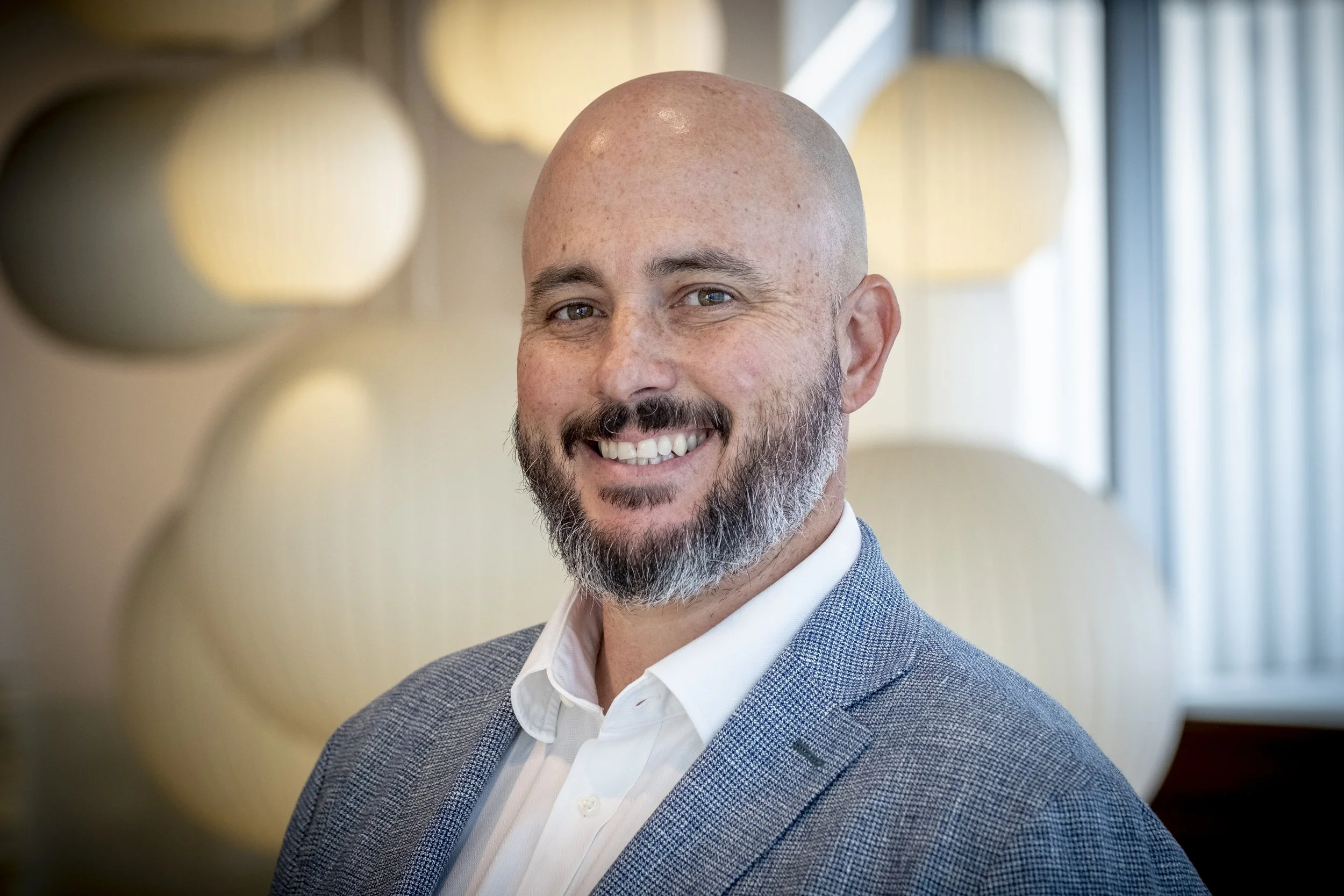A smiling man with a beard and bald head wearing a light gray blazer and a white shirt, standing in a well-lit indoor setting with blurred circular and rectangular window shapes in the background.