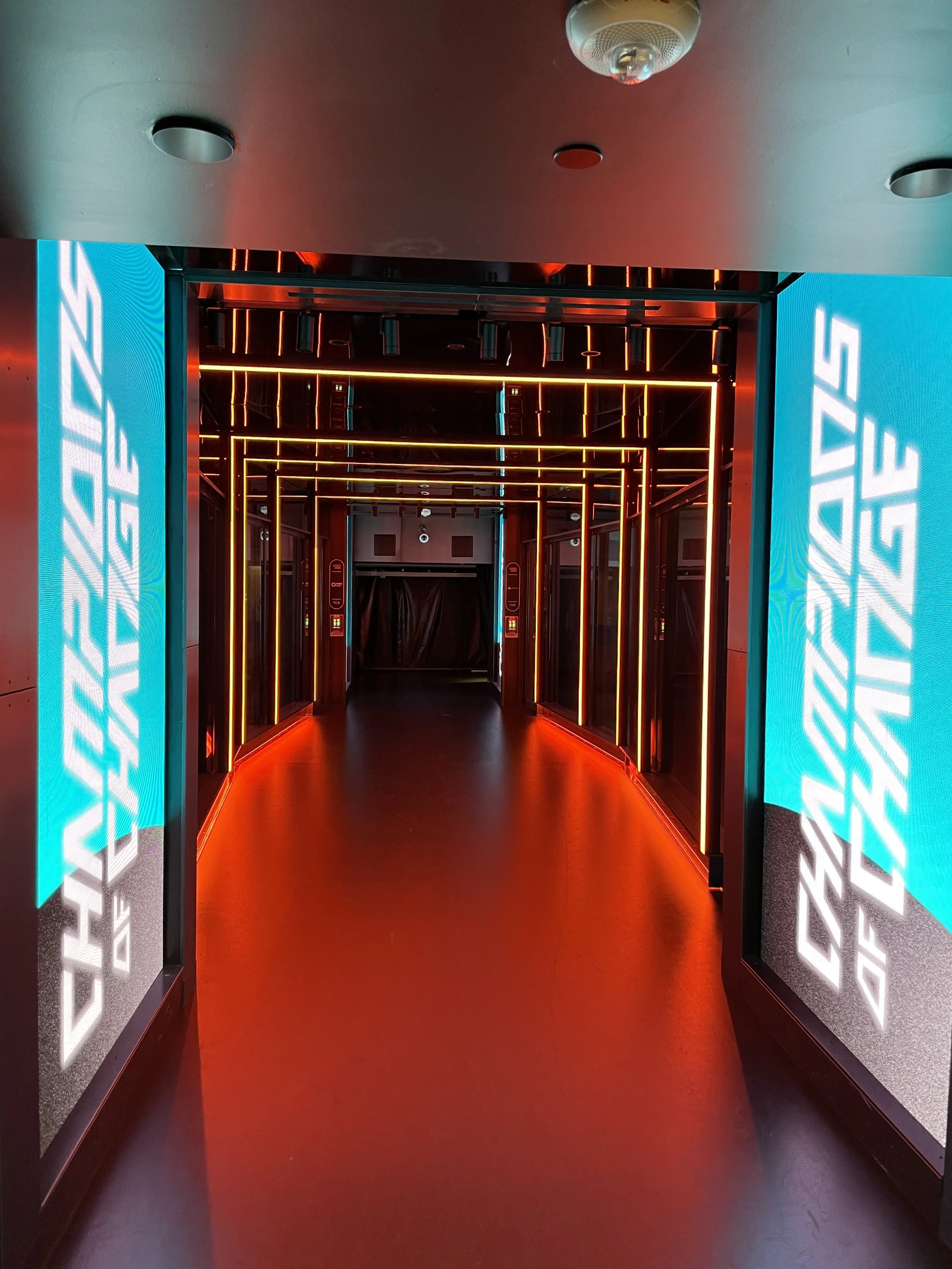 A futuristic hallway with red and orange neon lights outlining the ceiling and walls, leading to a dark doorway at the end.