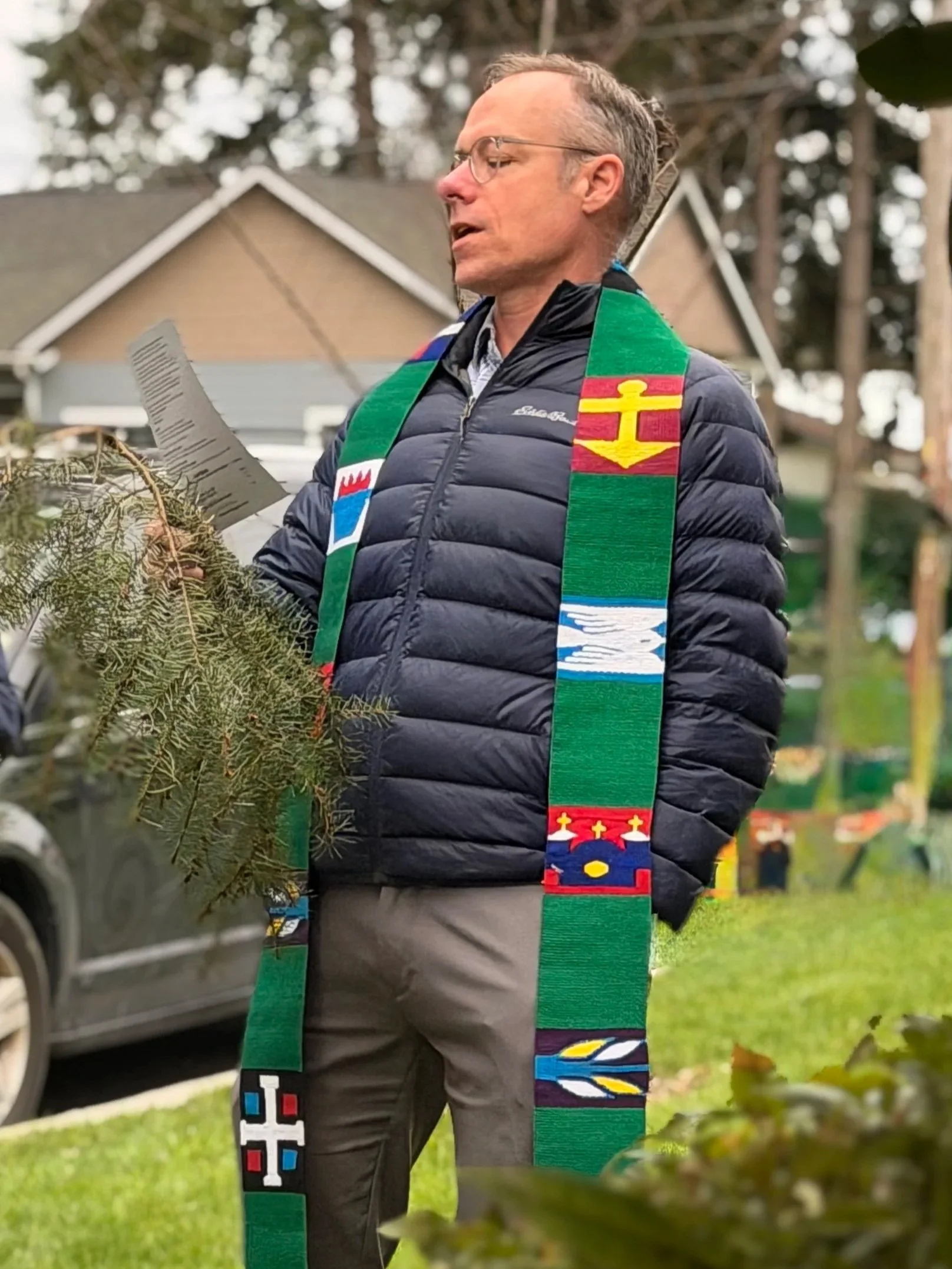 A Presbyterian minister wearing glasses and a dark puffer jacket with a colorful stole standing outdoors near a church and trees, holding a church bulletin.