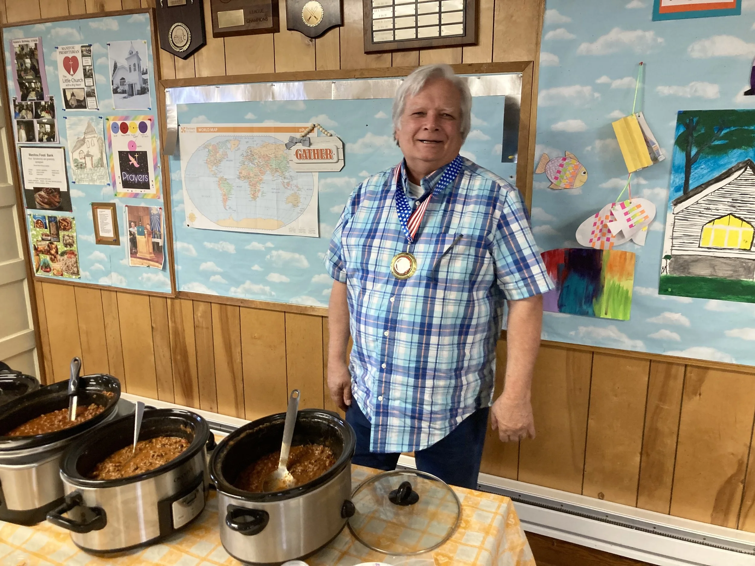 A man smiling and wearing a plaid shirt with a medal around his neck, standing behind a table with slow cooker dishes in a room decorated with a cloud sky mural, world map, and various posters.