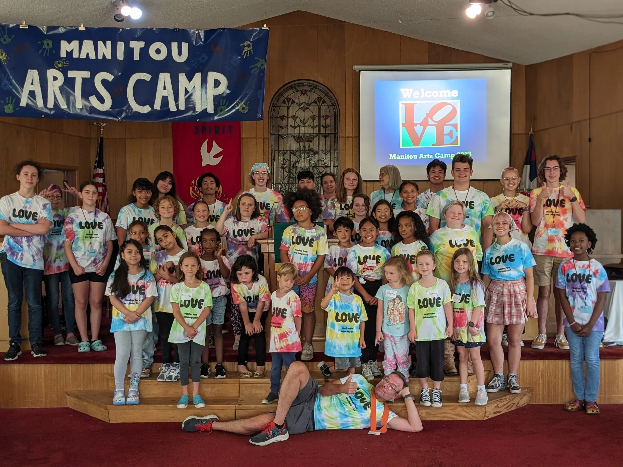 Group of children and adults on stage at Manitou Arts Camp, some wearing colorful tie-dye shirts with 'LOVE' written on them, in a wood-paneled room with banners and a projector screen.