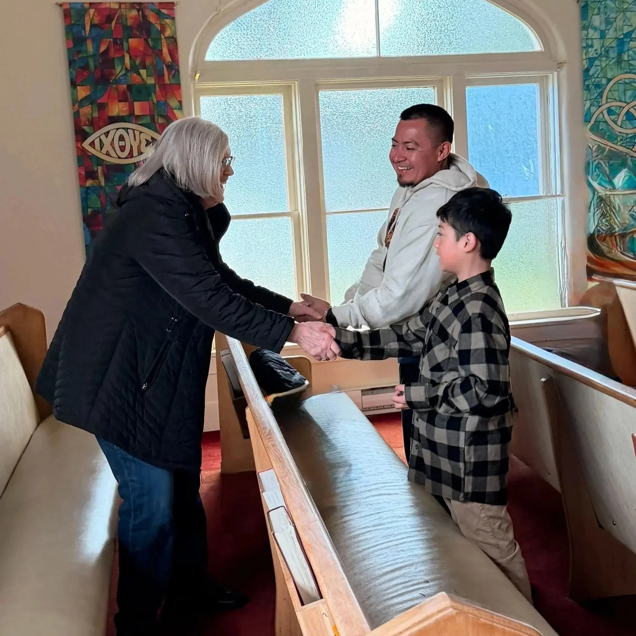 A woman and a father shake hands and smile at each other, along with the man's son between church pews inside a church with glass windows and colorful artwork.