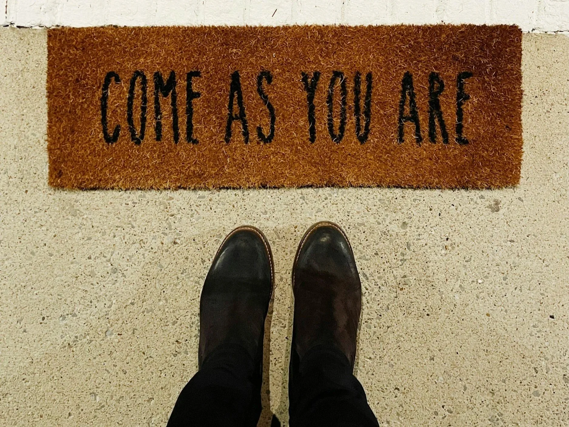View of a doormat that says, 'Come as you are,' with brown boots worn by a person visible at the bottom of the image, standing on a textured floor.