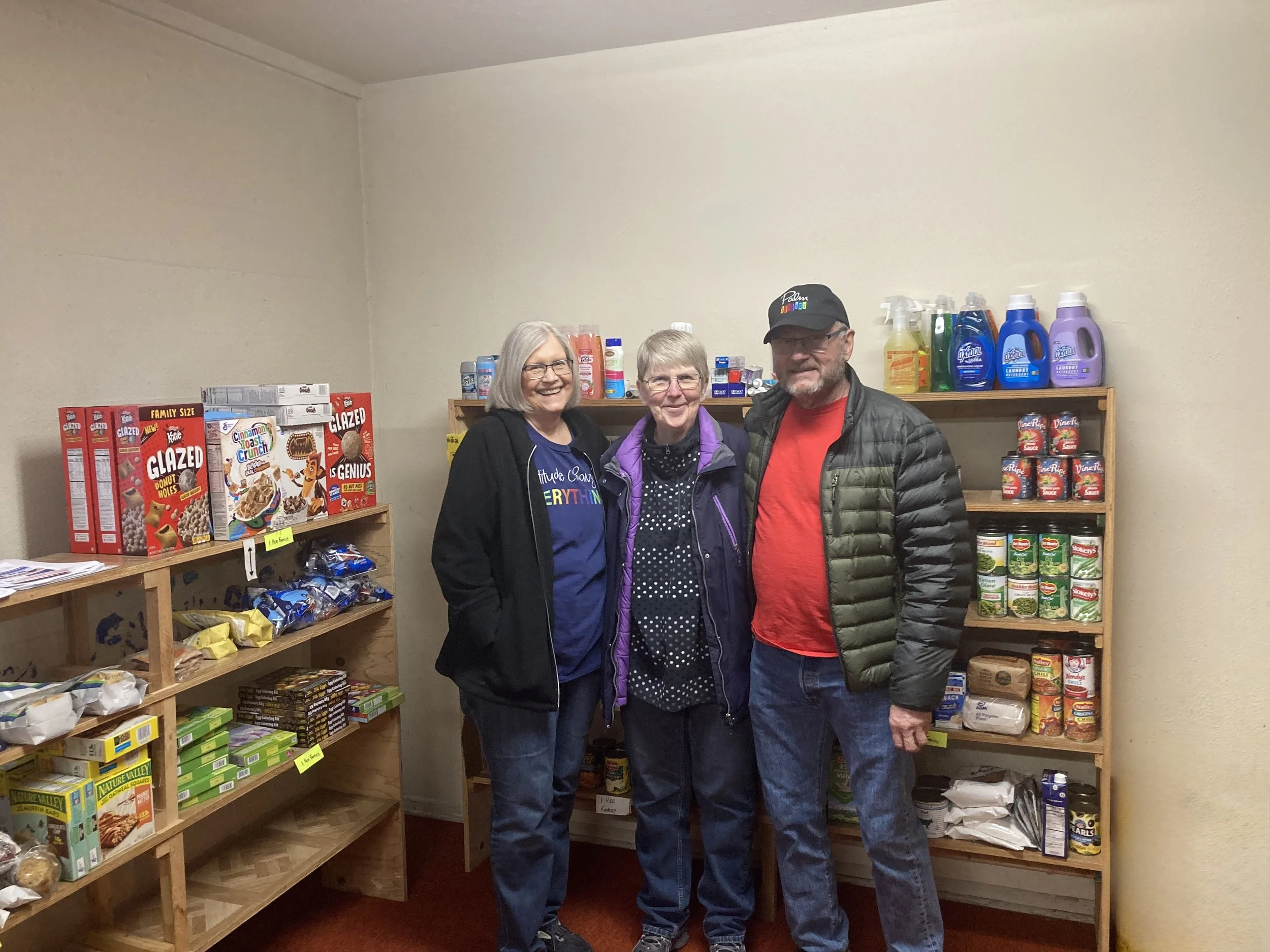 Three people standing inside a room with wooden shelves filled with food items like cereal, snack packs, and canned goods. They are smiling at the camera.