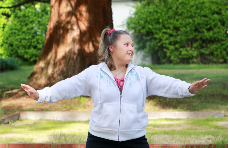A young woman with Down syndrome smiling, with a white zippered jacket, ponytail, and pink hair accessories standing outdoors with her arms extended to the sides in front of a large tree and green bushes.