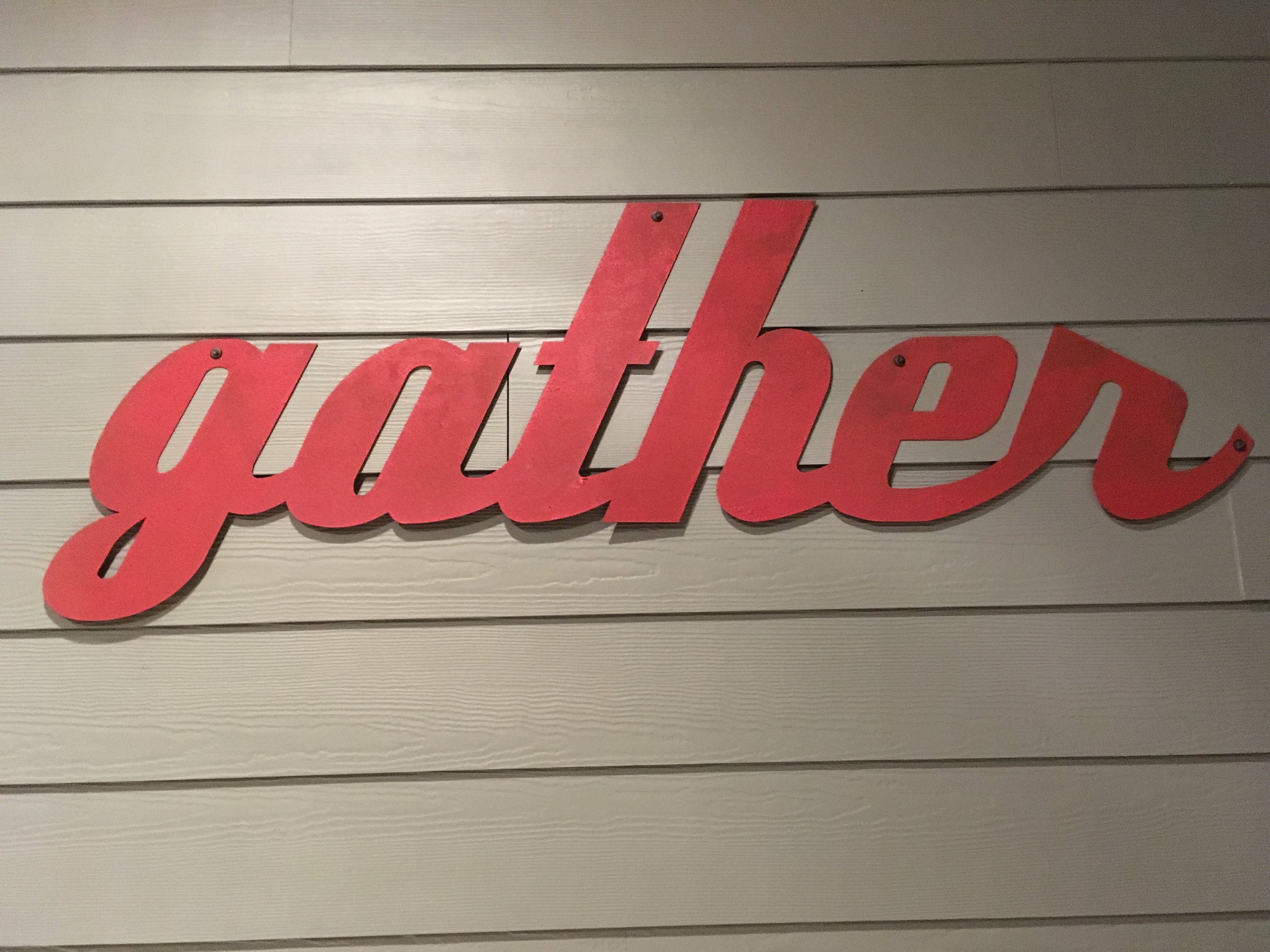 Red cursive letter sign that reads "gather" mounted on wooden panel wall.