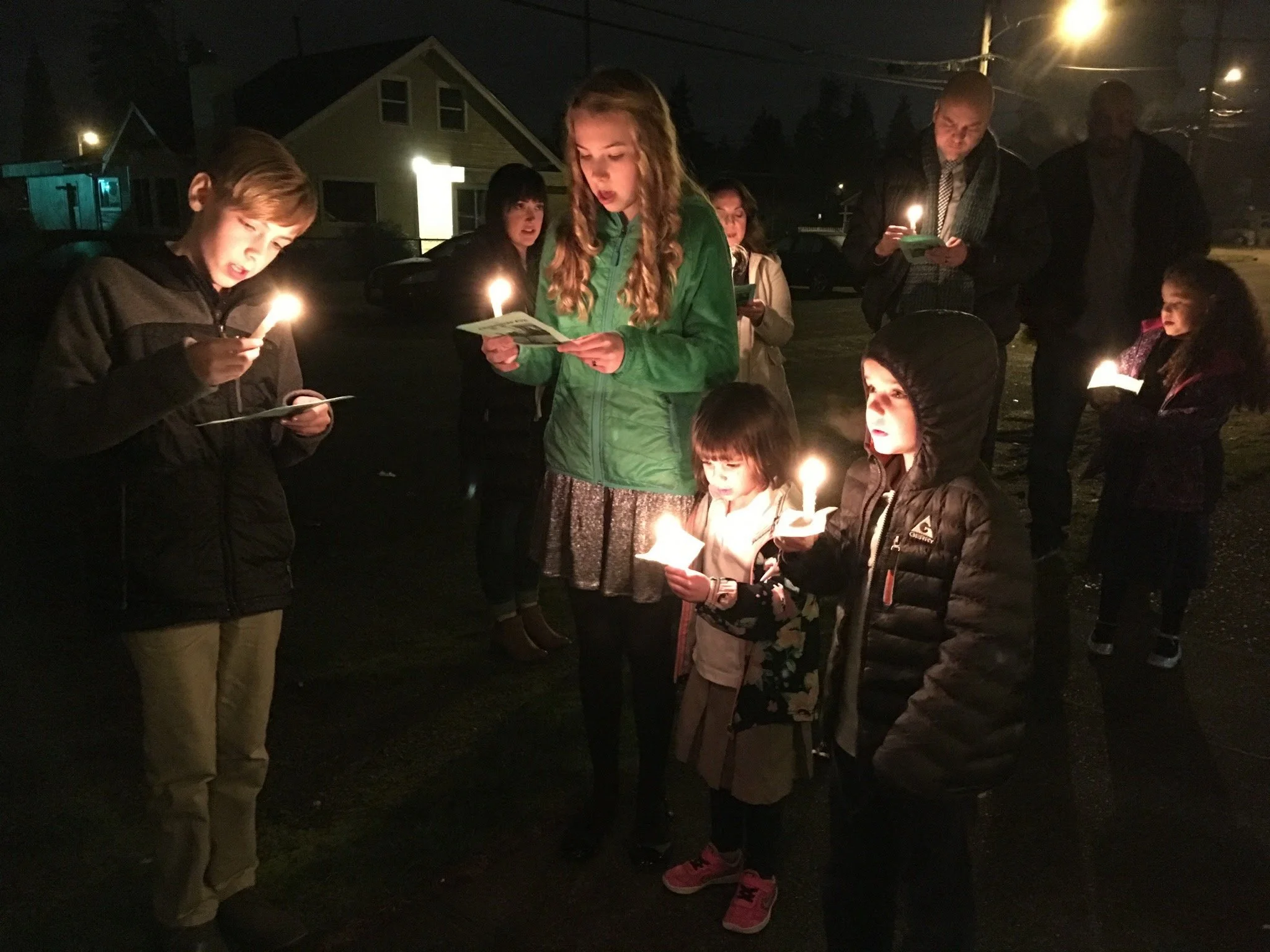 A group of children and a few adults gathered outdoors at night, holding candles and reading from papers, likely during a candlelit vigil or ceremony.