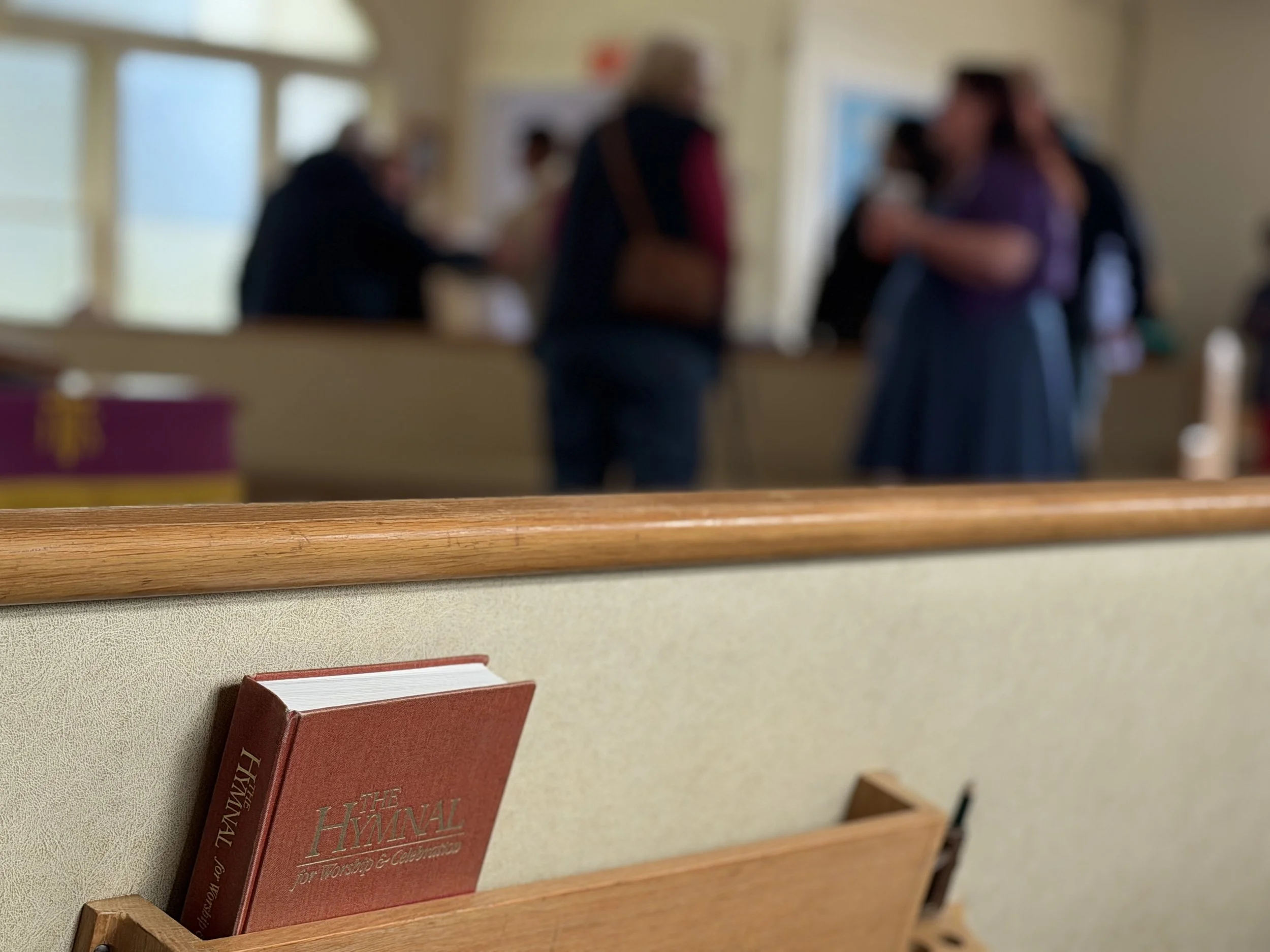 A red book titled "Hymnal" placed on wooden rack behind a pew, with a blurred background of people in a church.