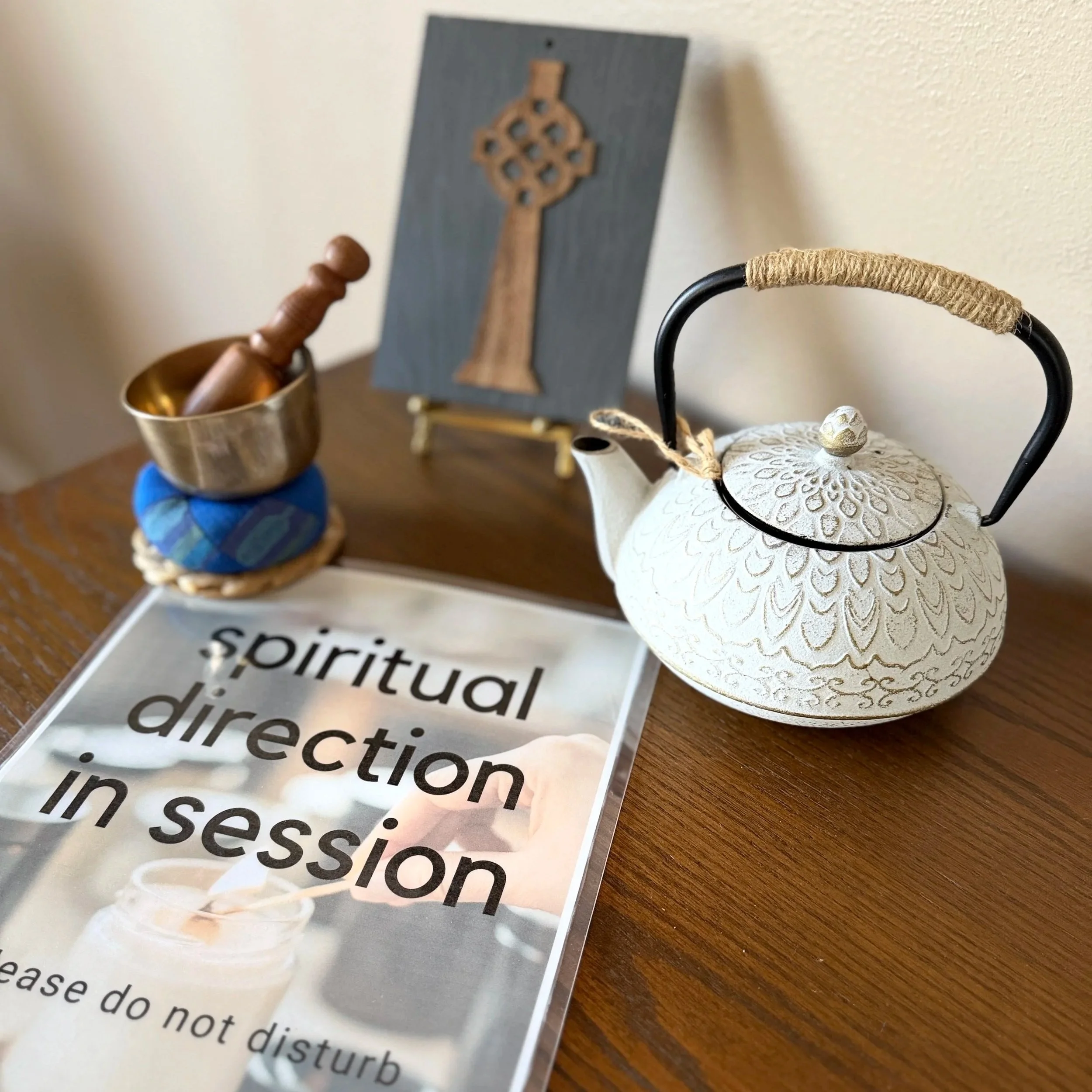 Close-up of a table display featuring a decorative white teapot, a small sign with the word 'spiritual' printed multiple times, a wooden stand with a Nordic-inspired ornament, and a small brass bowl with a spoon.