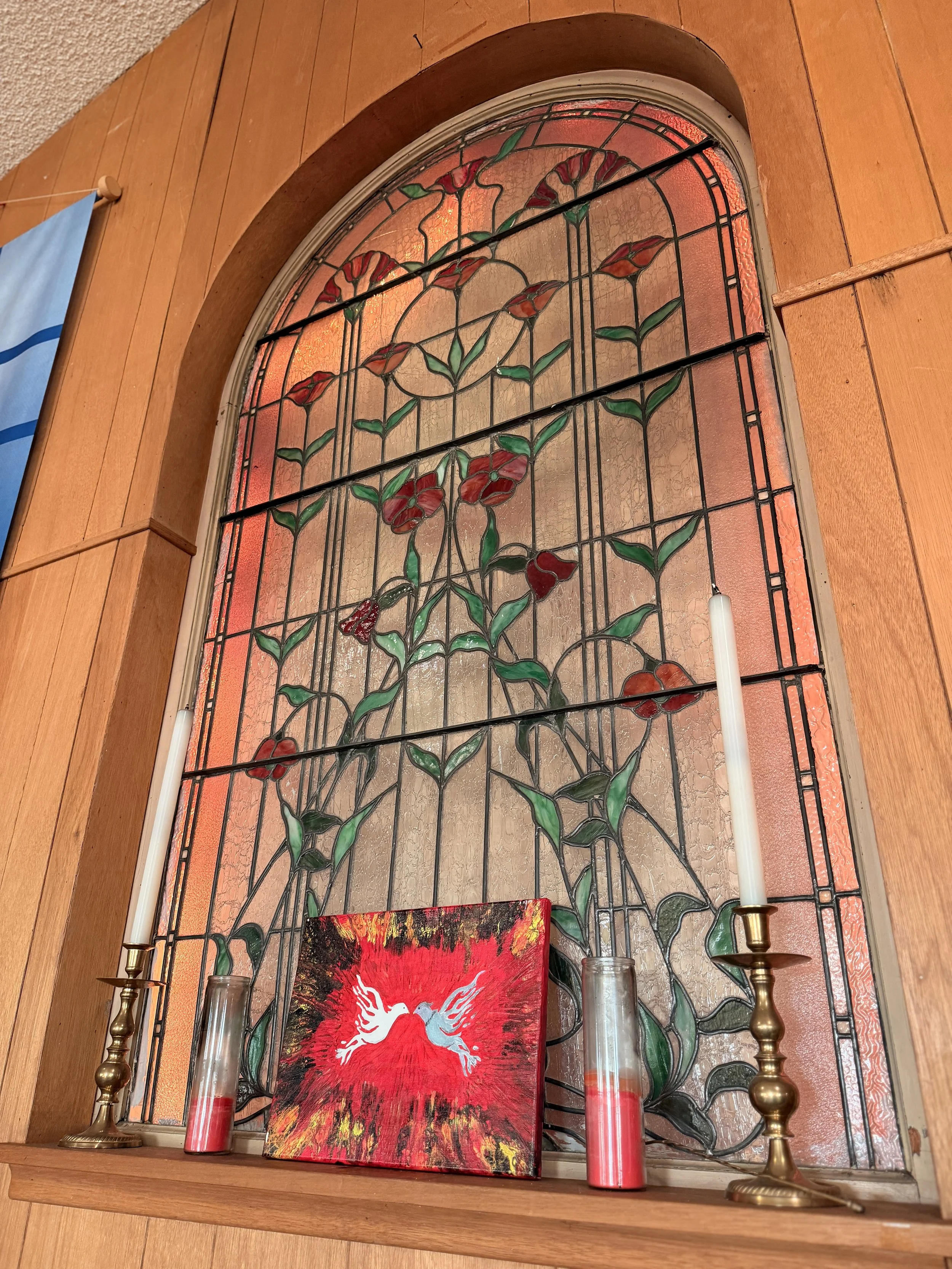 A stained glass window with a floral design, flanked by two brass candlesticks with white candles, and a red painting with a white dove and wings at the bottom.
