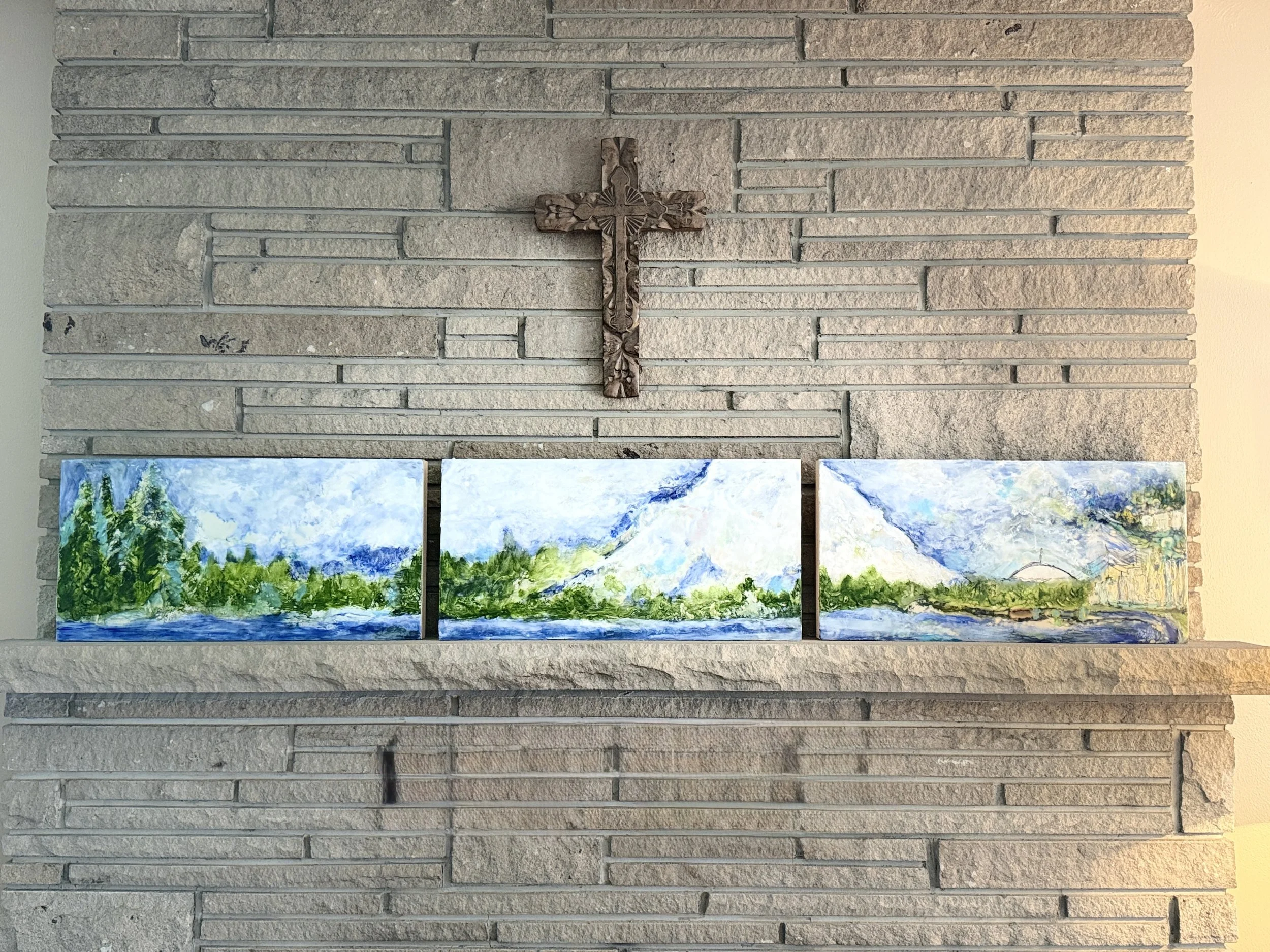 A decorative wooden cross hanging on a gray brick wall, with abstract landscape paintings at the bottom.