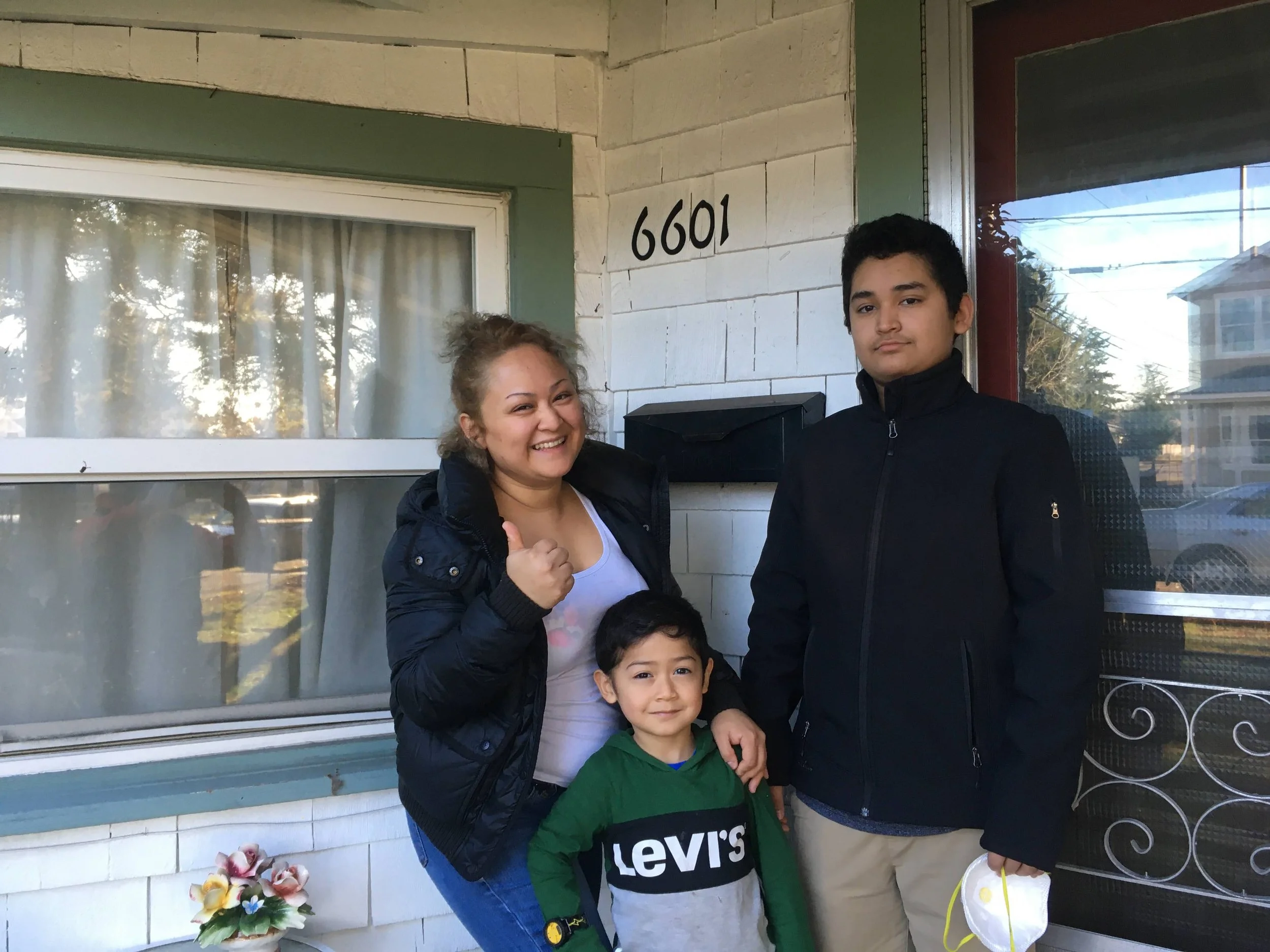 A group of three people, two children and a woman, stands outside a house with the address 6601, smiling and posing for the photo. The woman is giving a thumbs up, and they are in front of a window and door, with flowers on the porch.