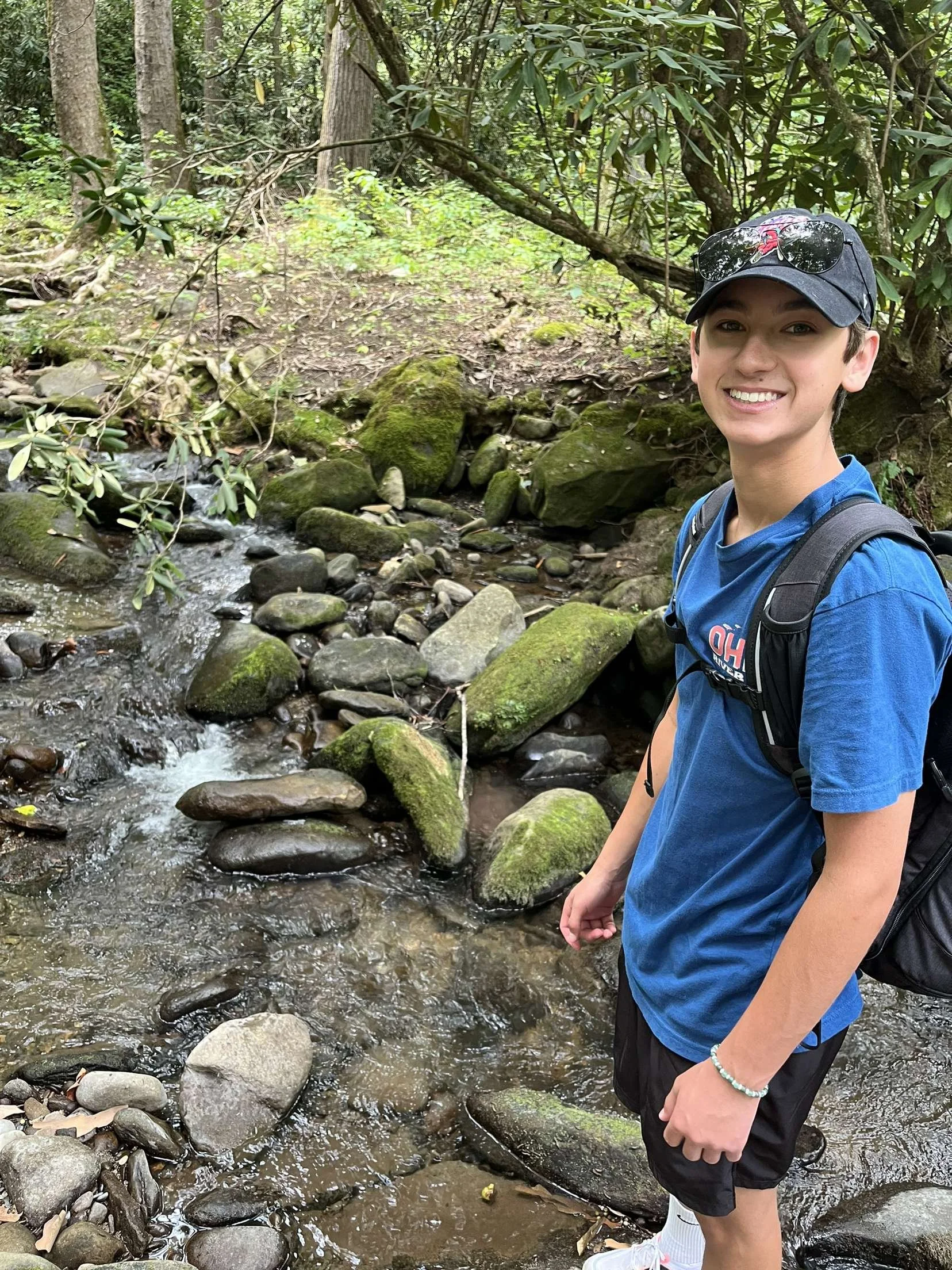 A young man smiling while standing in a shallow mountain stream surrounded by rocks and green forest.