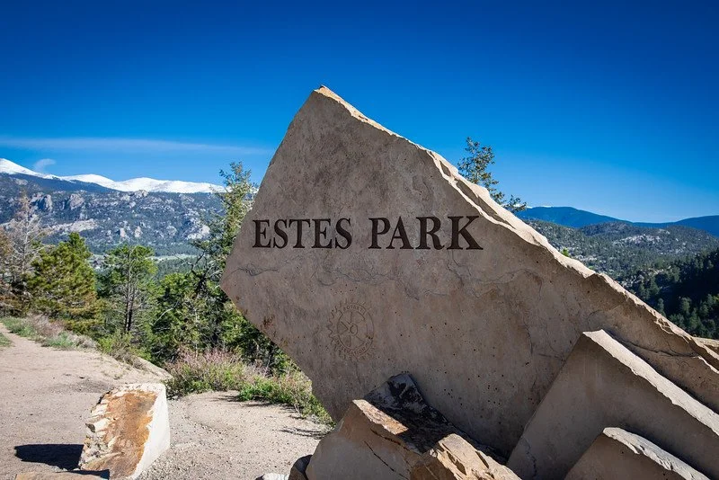 A large, tilted stone sign with the words 'Estes Park' engraved on it, set against a mountain landscape with a bright blue sky.