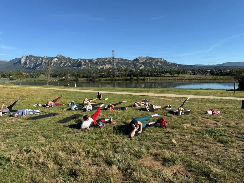 A group of people practicing yoga outdoors on a grassy field next to a lake, with mountains in the background and a clear blue sky overhead.