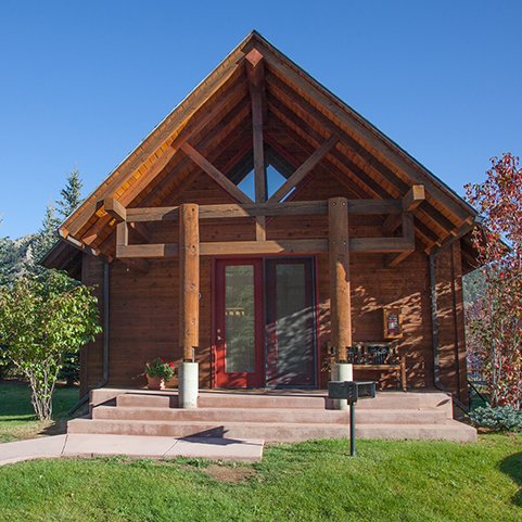 A small wooden house with a pitched roof, front porch, and red door, surrounded by a green lawn and trees.