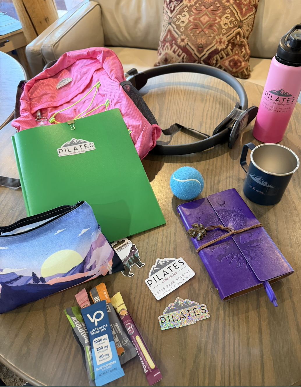 Table with various items related to Pilates, including a pink backpack, green folder with a Pilates logo, black and silver headphones, pink water bottle, blue tennis ball, purple journal, black mug with a mountain logo, patch with 'Pilates' text, energy drink tubes, and stickers, set against a background with a cushion and a cat sleeping.
