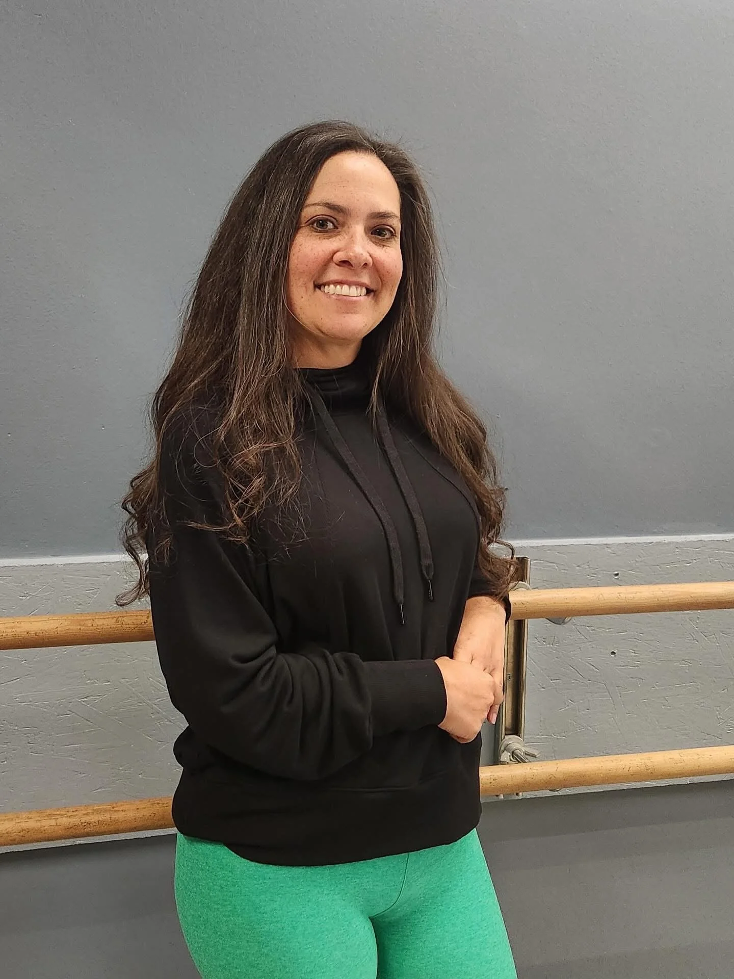 A woman with long brown hair and a smile, wearing a black hoodie and green leggings, standing in front of a ballet barre against a gray wall.