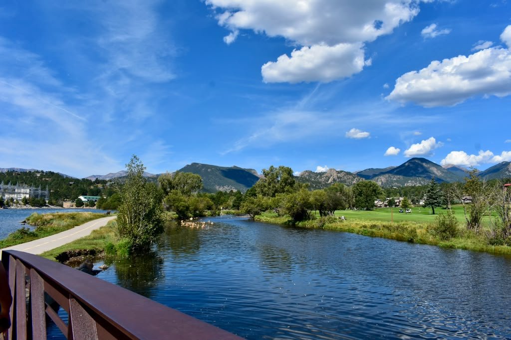 Scenic view of a river with a walking path on the left, surrounded by green trees and grass, with mountains and a partly cloudy blue sky in the background.