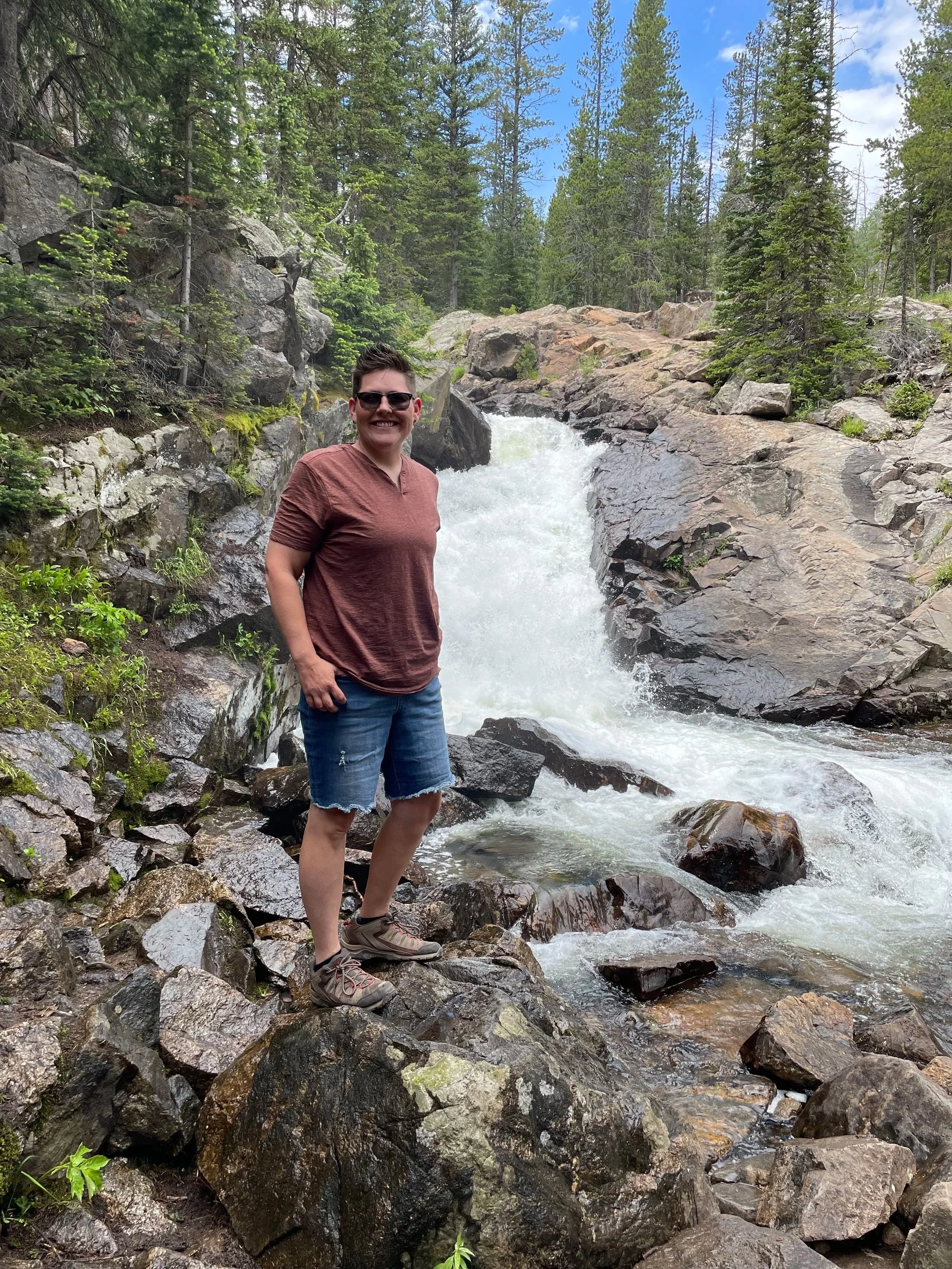 A woman wearing sunglasses, a maroon T-shirt, and denim shorts standing on rocks beside a waterfall in a forested area.
