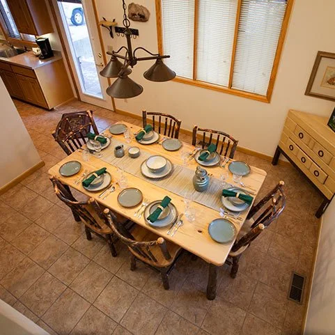 A dining room with a wooden table set for eight people, featuring plates, napkins, and utensils, surrounded by wooden chairs, with a chandelier hanging overhead and a sideboard against the wall.
