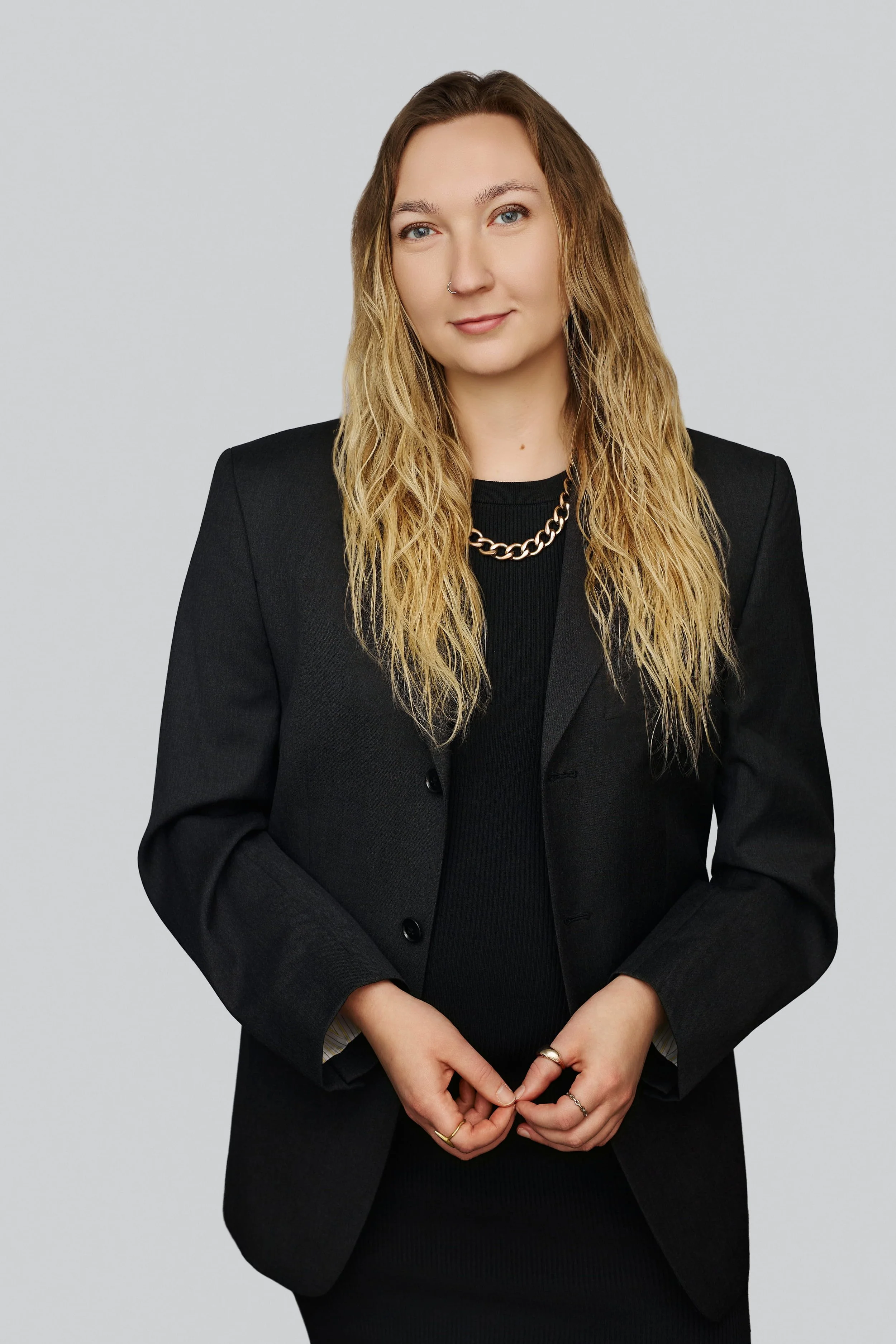 A woman with long, wavy blonde hair and a nose piercing wearing a black blazer, black top, a gold chain necklace, and rings on her fingers, standing against a plain gray background.