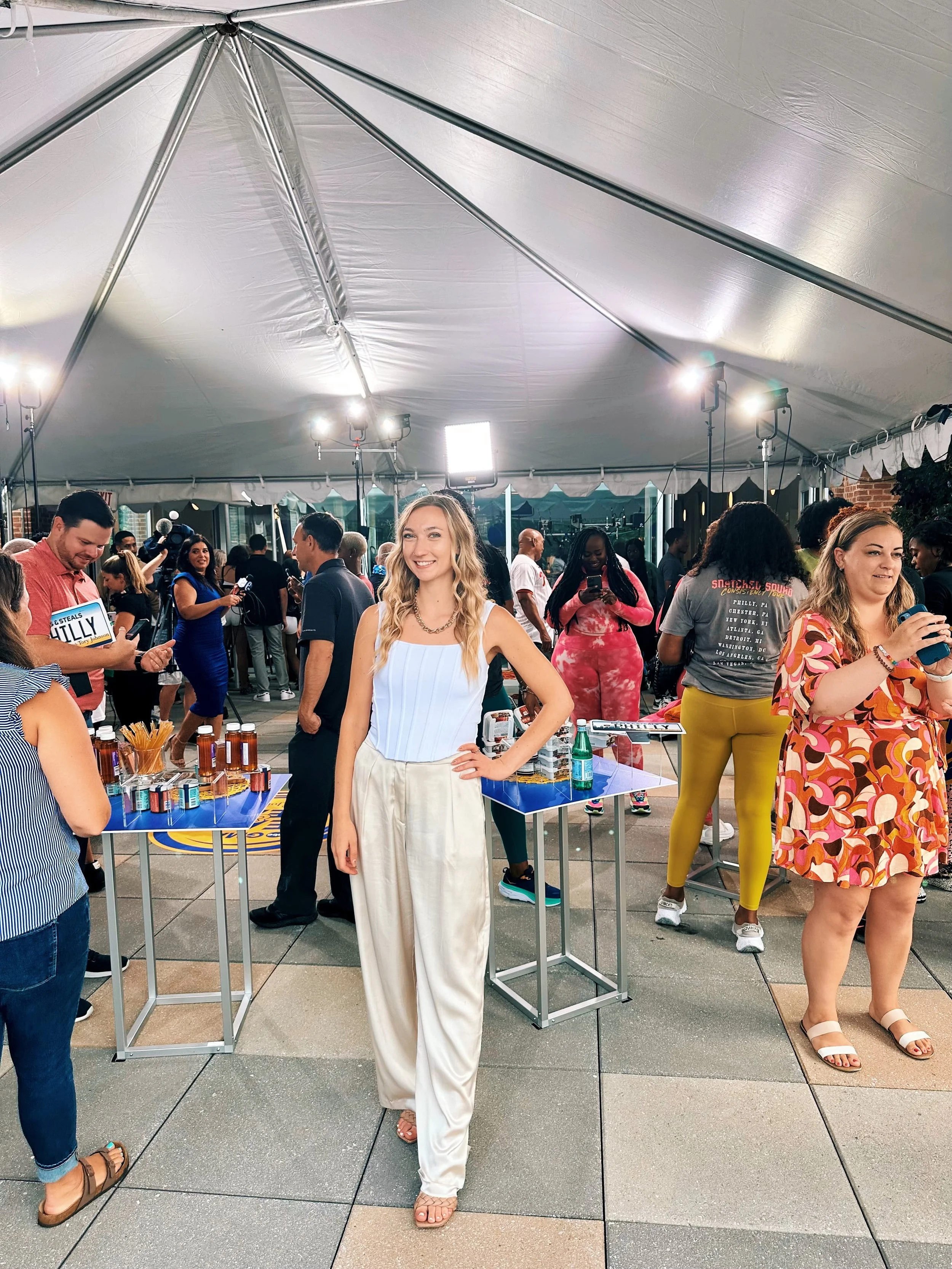 A woman with long blonde hair, wearing a white sleeveless top and cream wide-leg pants, stands smiling at an outdoor event under a large white tent. There are tables with bottles and products, and several people mingling and taking photos in the background.