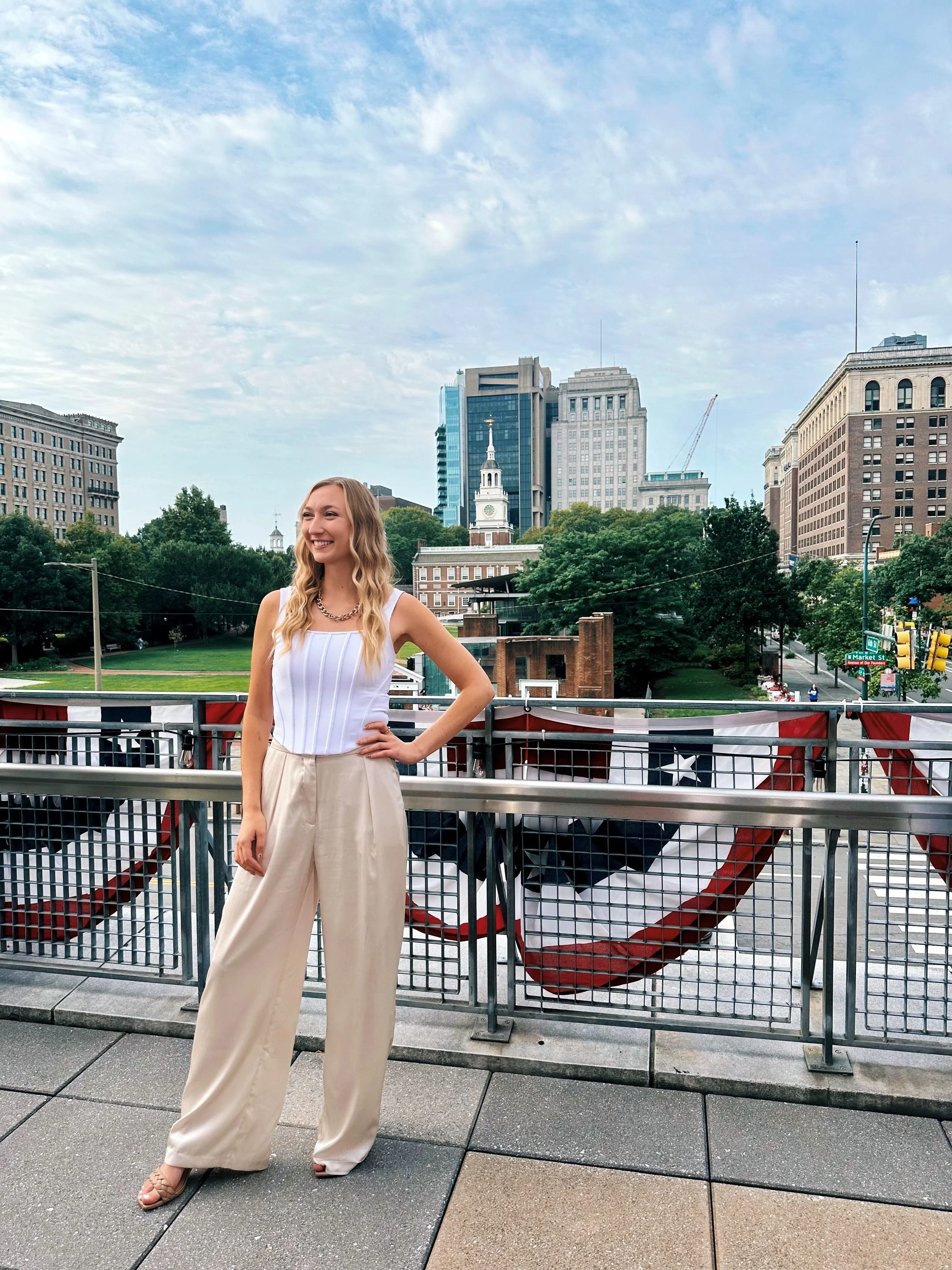 A woman smiling and posing on a balcony decorated with a large American flag. City buildings and trees are visible in the background.