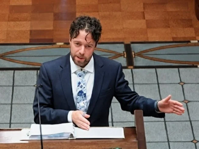 A man in a dark suit, white shirt, and floral tie, speaking or presenting at a wooden desk in a formal setting. He has dark, curly hair and a beard, and appears to be gesturing with his right hand.