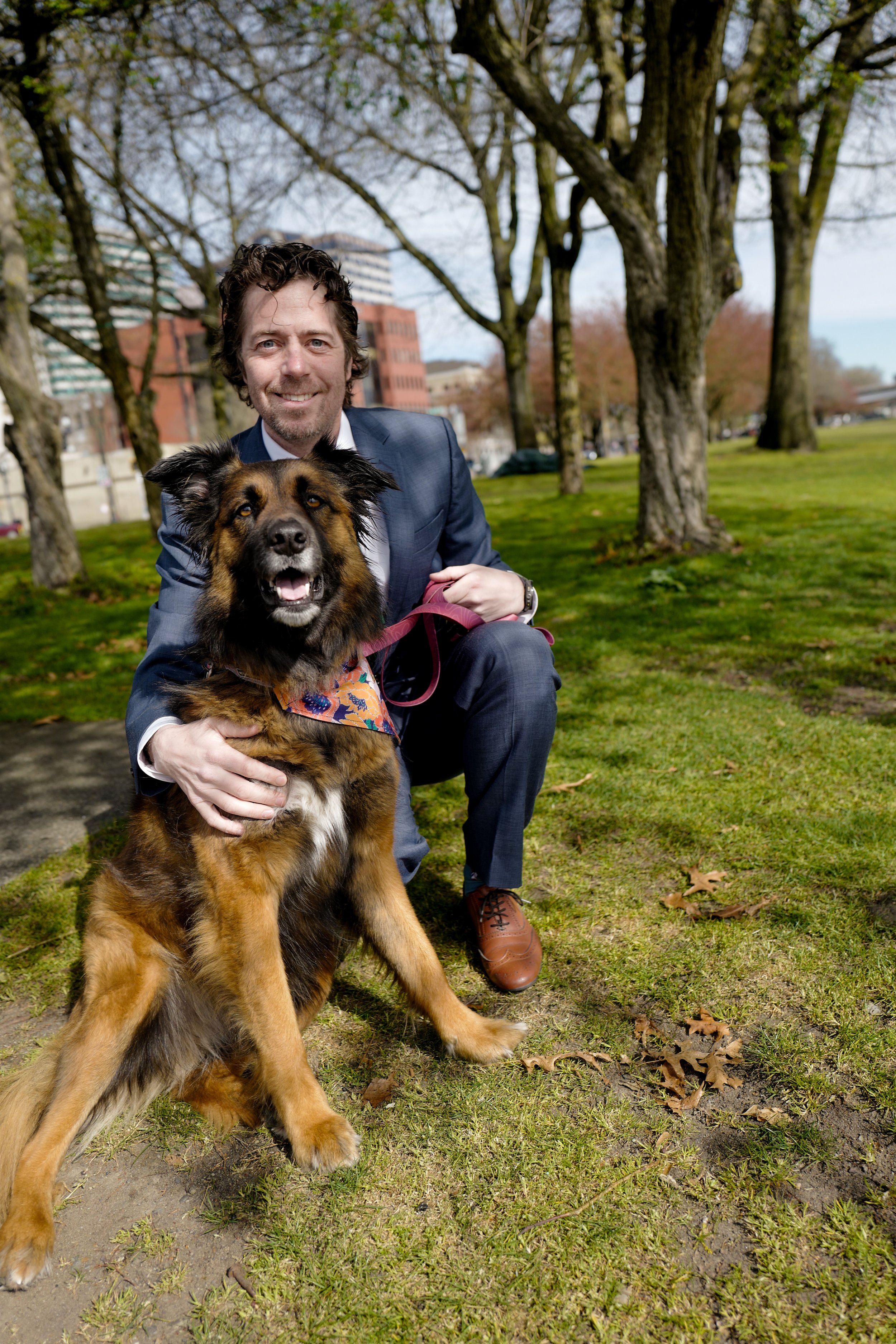 A man in a suit crouching beside a happy large dog with a colorful bandana outdoors in a park with trees and buildings in the background.