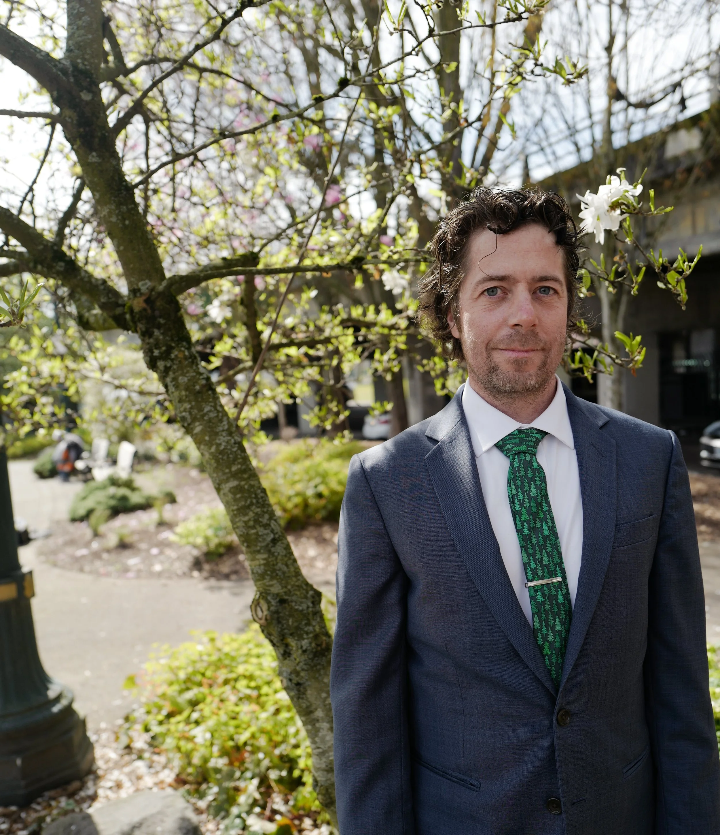 A man in a suit and tie standing outdoors in front of a flowering tree.