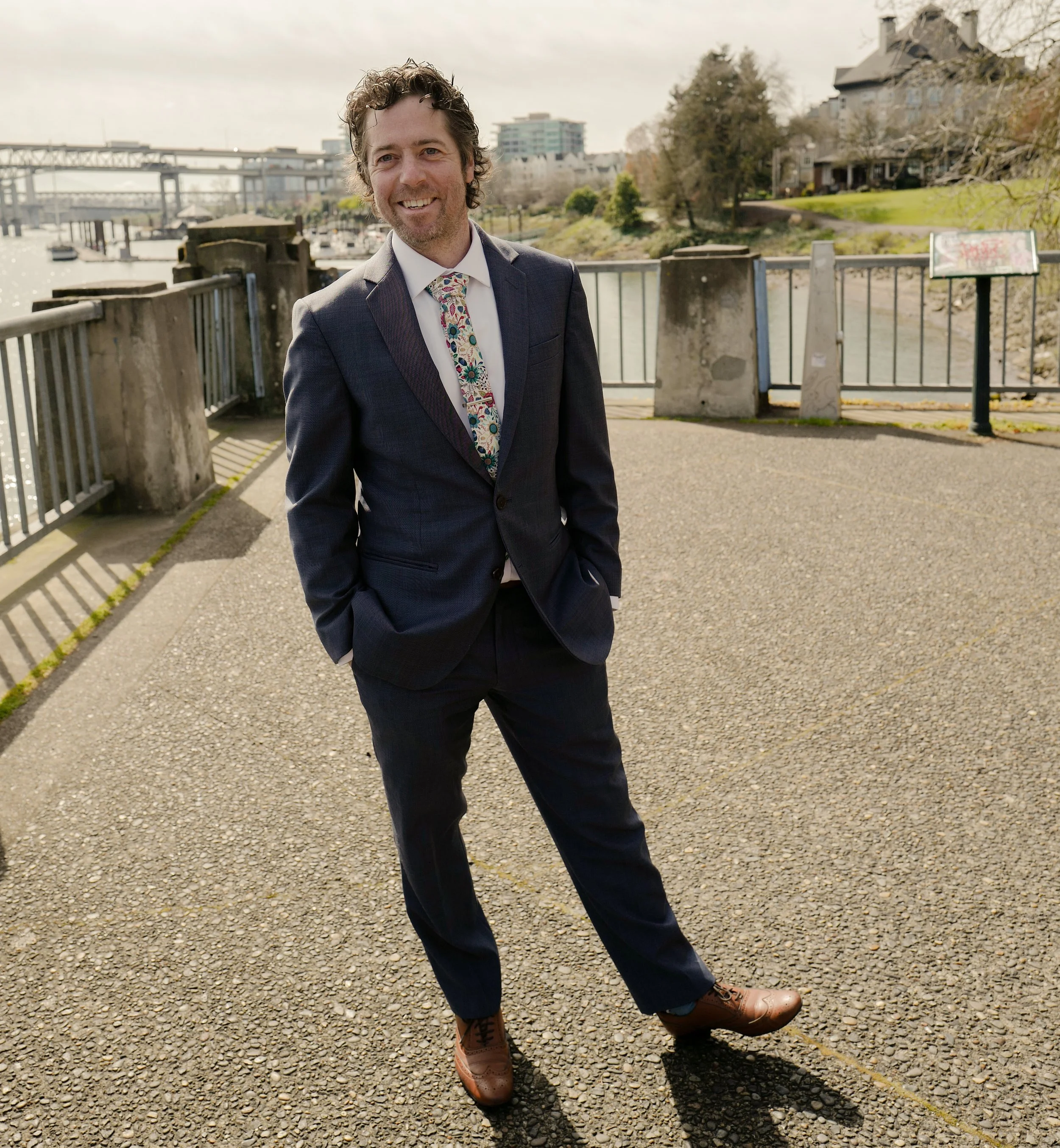 A man wearing a dark suit, white shirt, and floral tie standing outdoors on a paved walkway near a river, with buildings and a bridge in the background.