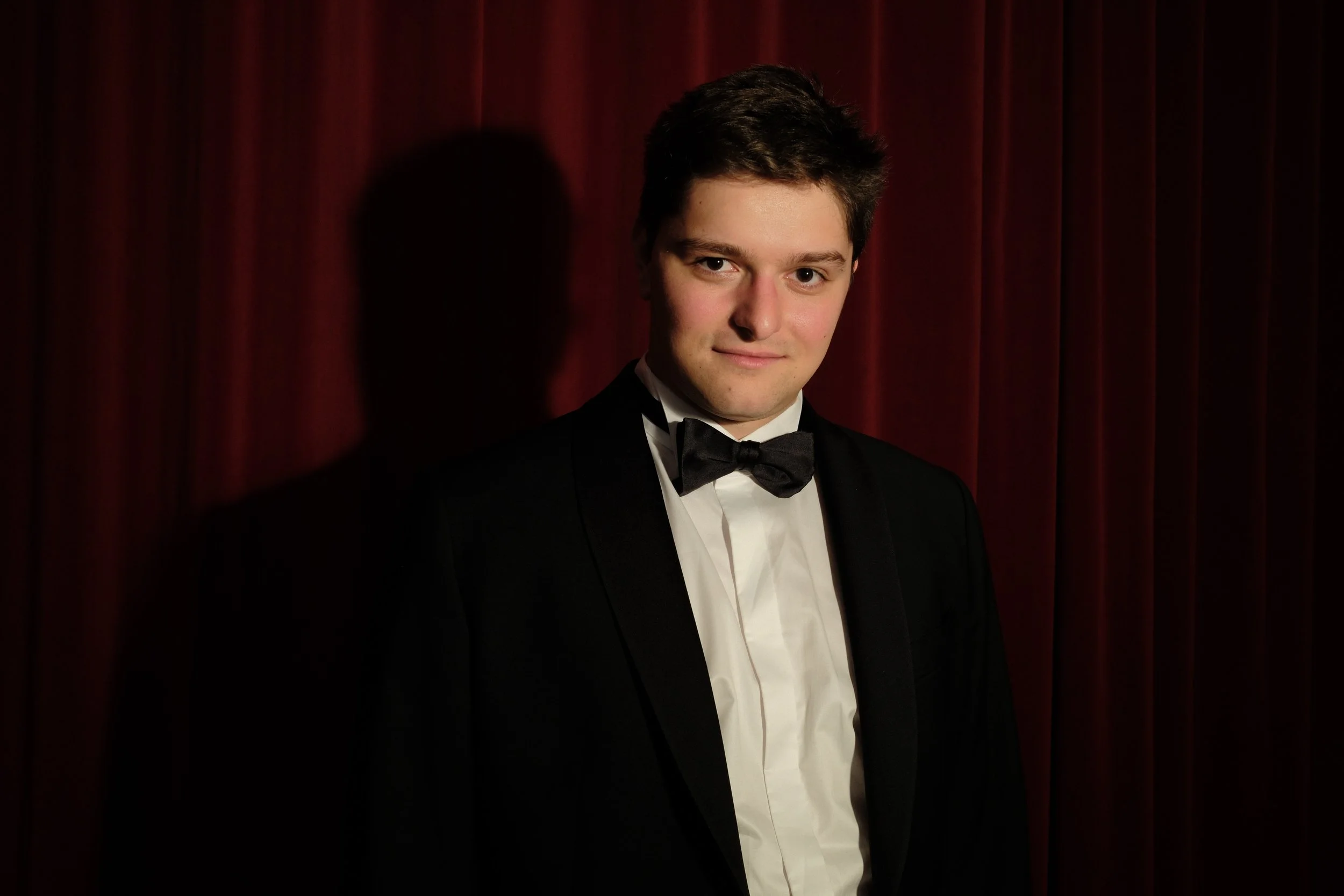 Young man in tuxedo with bow tie standing in front of red velvet curtain.