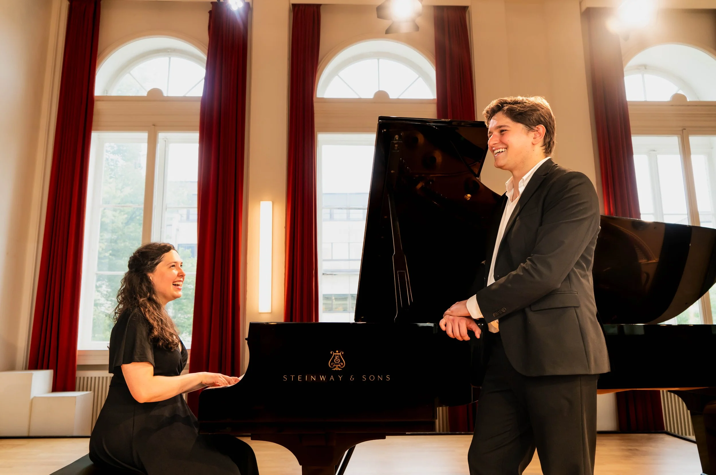 A woman sitting at a black grand piano, smiling and looking up at a man standing beside her, who is dressed in a dark suit and smiling back. They are in a bright room with large arched windows and red curtains, and a person is visible sitting behind the piano. The scene appears to be a musical performance or rehearsal.