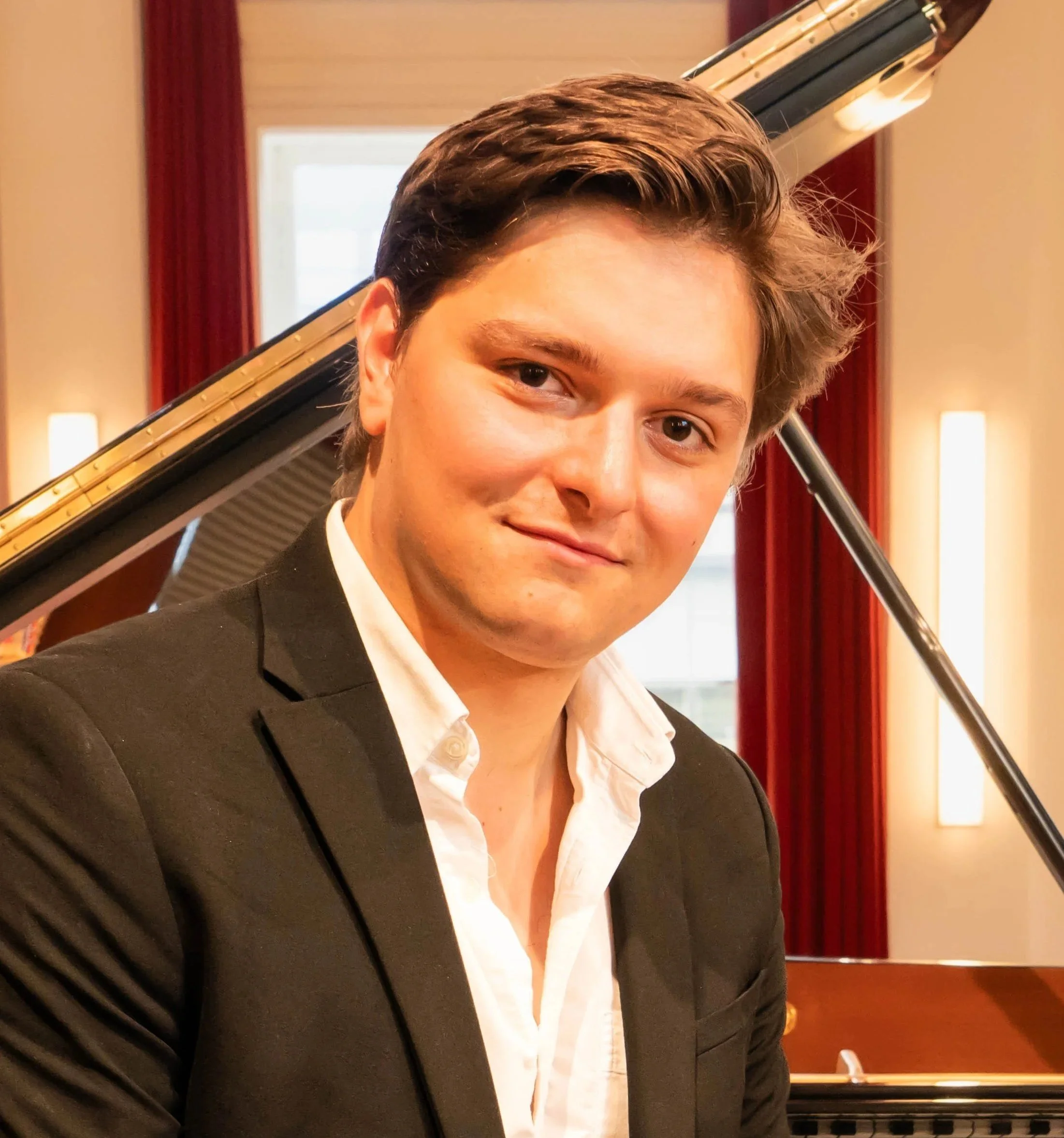 Close-up of a young man with brown hair and light skin, wearing a black tuxedo and white shirt, sitting at a grand piano in a room with warm lighting and red curtains.